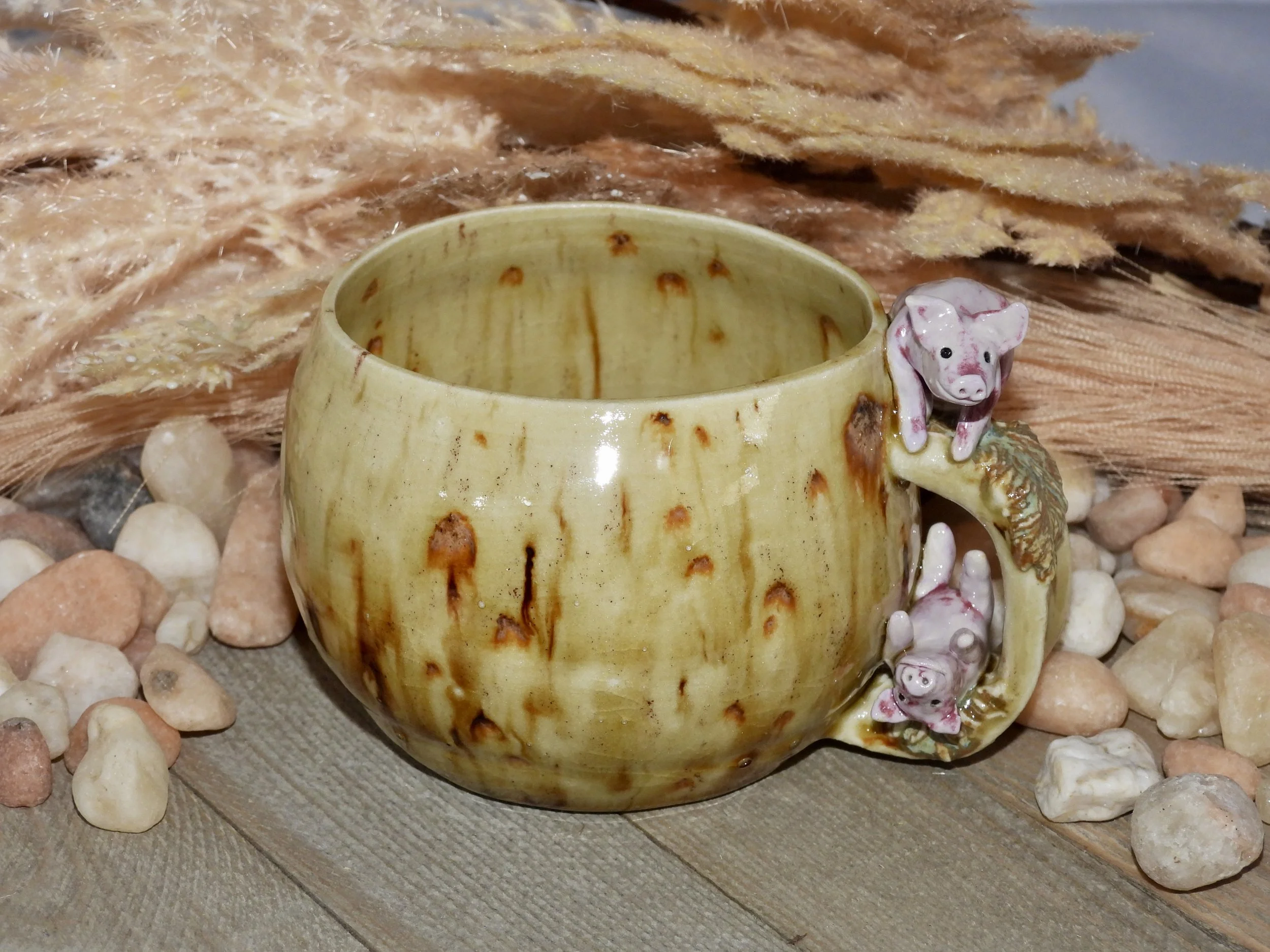 A ceramic mug decorated with a pig figure on the handle, placed on a wooden surface surrounded by small stones and beige dried grass.