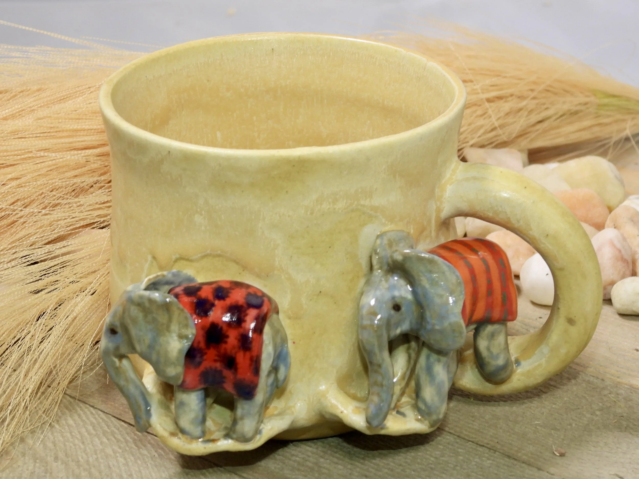 A yellow ceramic mug with two small elephant figurines attached, each wearing a red and orange garment, on a wooden surface surrounded by pebbles and dried grass.