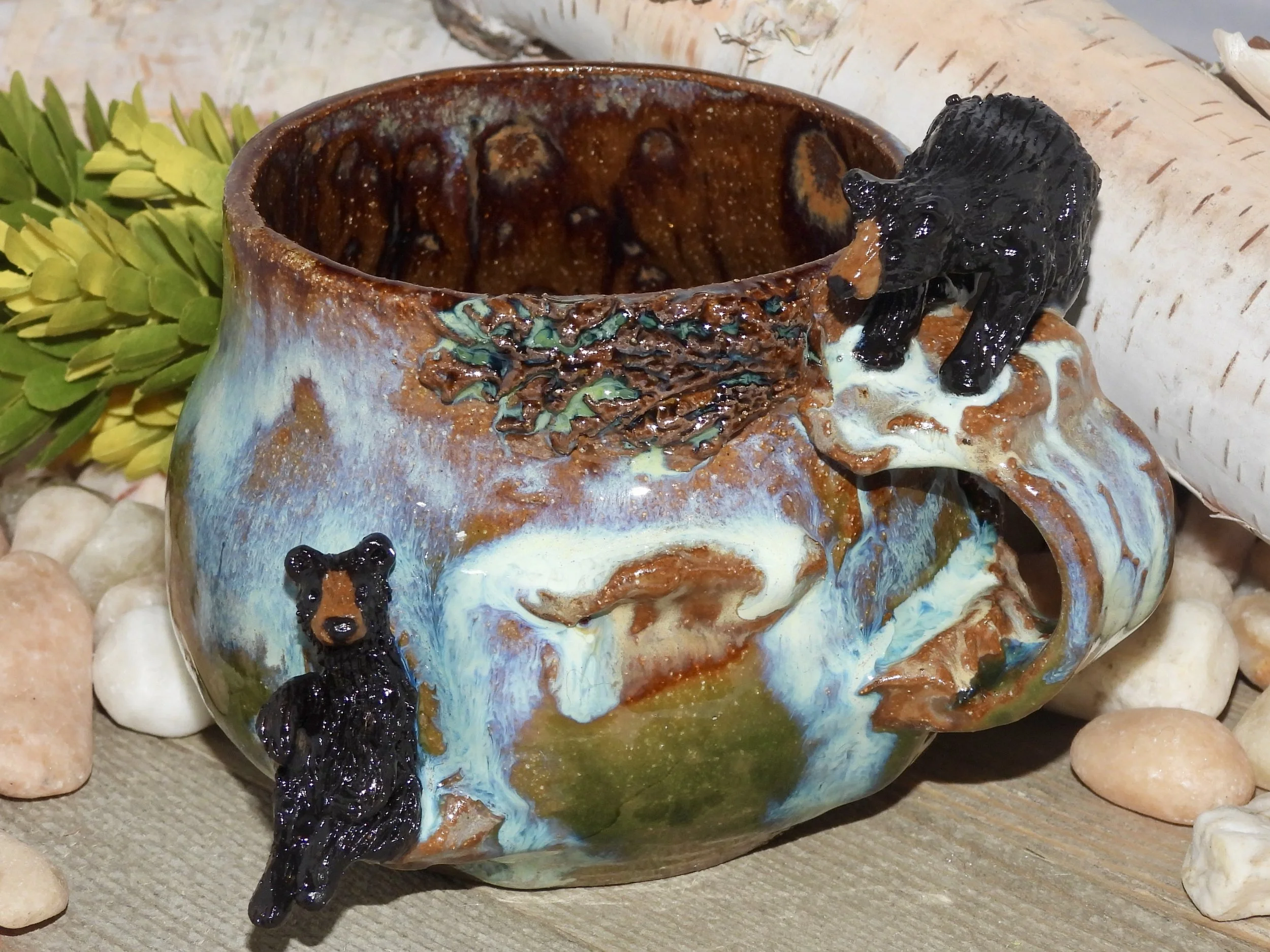 A ceramic mug with a mountain scene and two black bear figurines, one on top and one hanging from the mug's edge, surrounded by rocks and greenery.