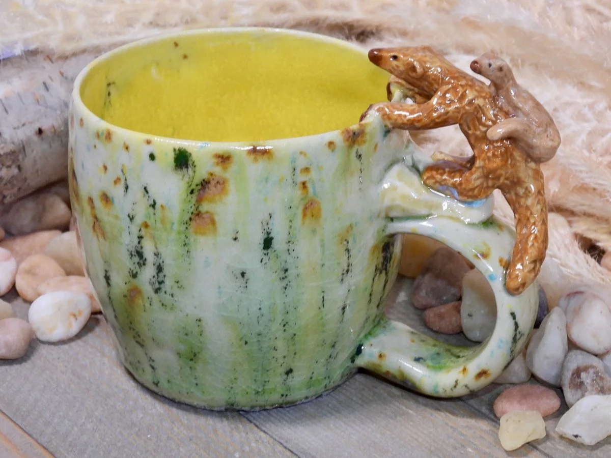 A ceramic mug with a crackled glaze finish and a frog sculpture attached to the handle, resting on a wooden surface surrounded by small rocks.