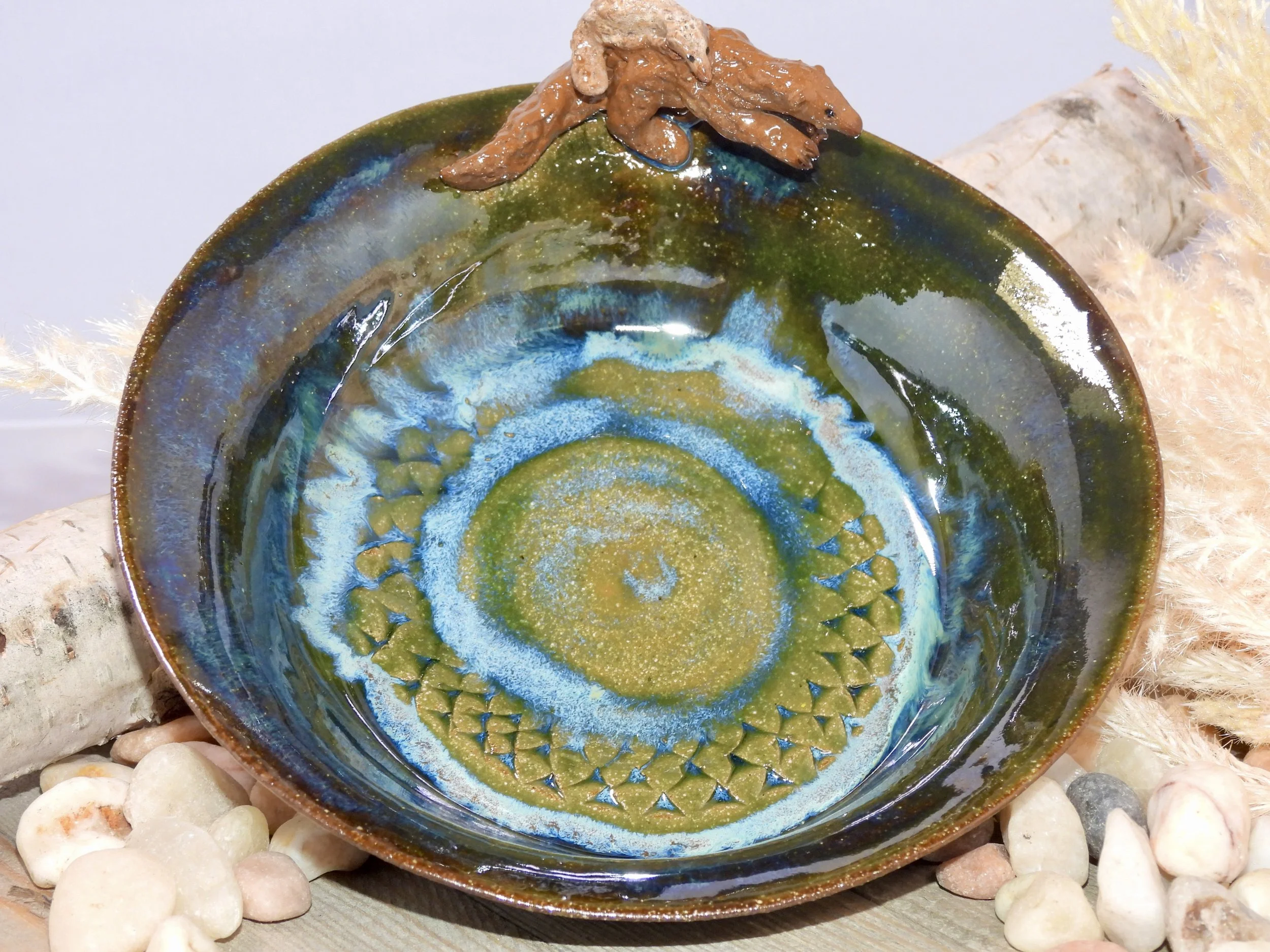 A colorful, glazed, ceramic bowl with a spiral pattern in shades of blue, green, and gold. The bowl is surrounded by small white stones and natural elements like a piece of wood and dried plants.