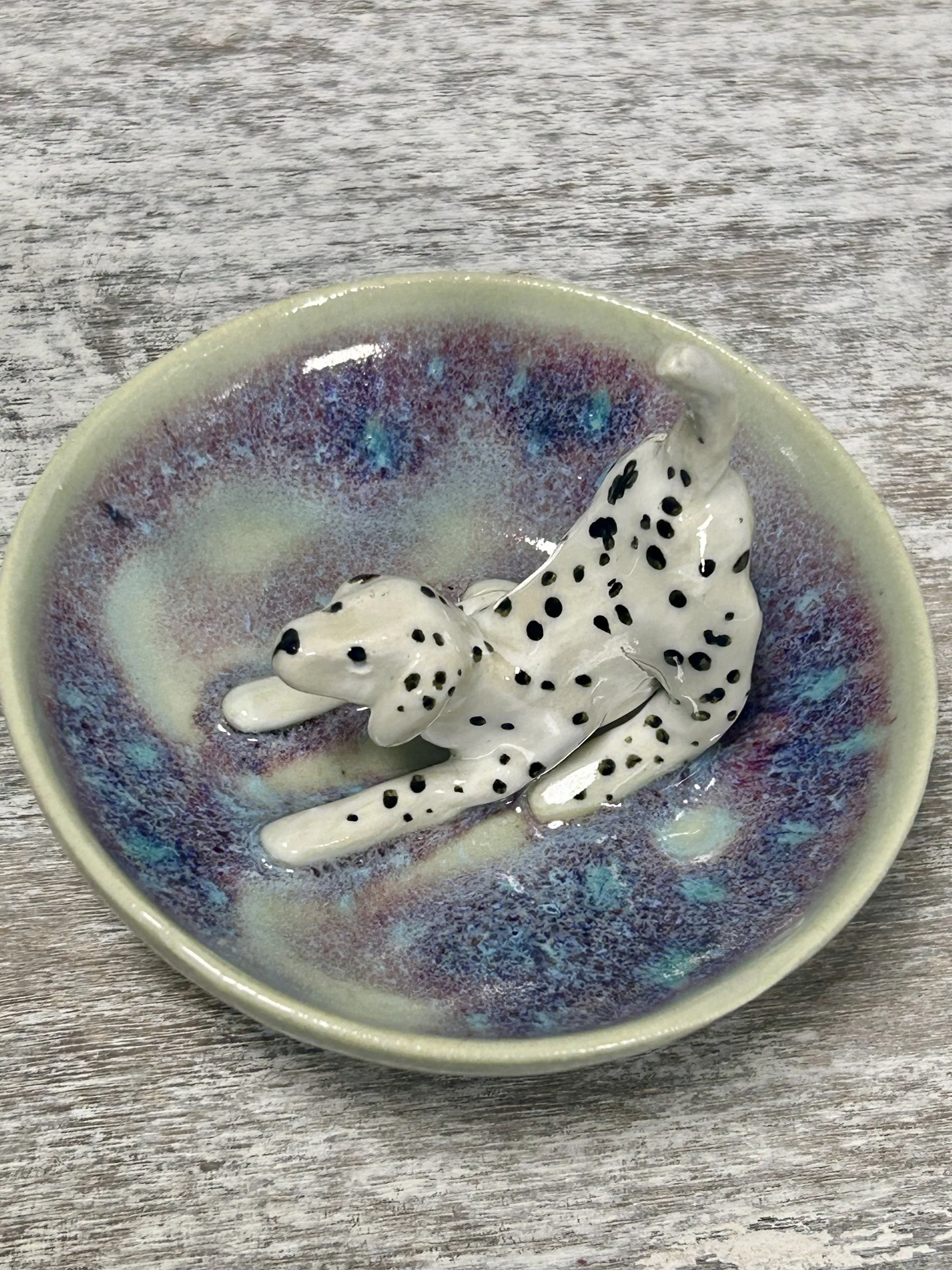 A ceramic dish shaped like a Dalmatian dog resting on a multicolored glazed bowl with an iridescent finish, placed on a wooden surface.