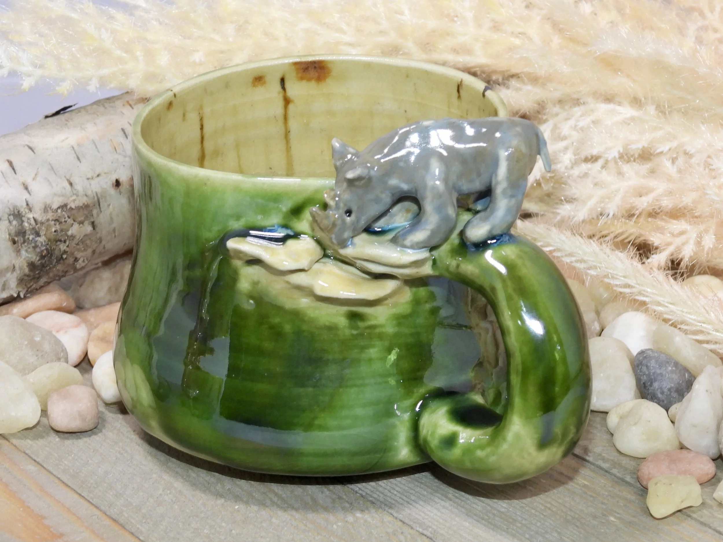 Ceramic mug with a green glaze and a small gray dog figurine on the rim, surrounded by rocks and soft feathers.