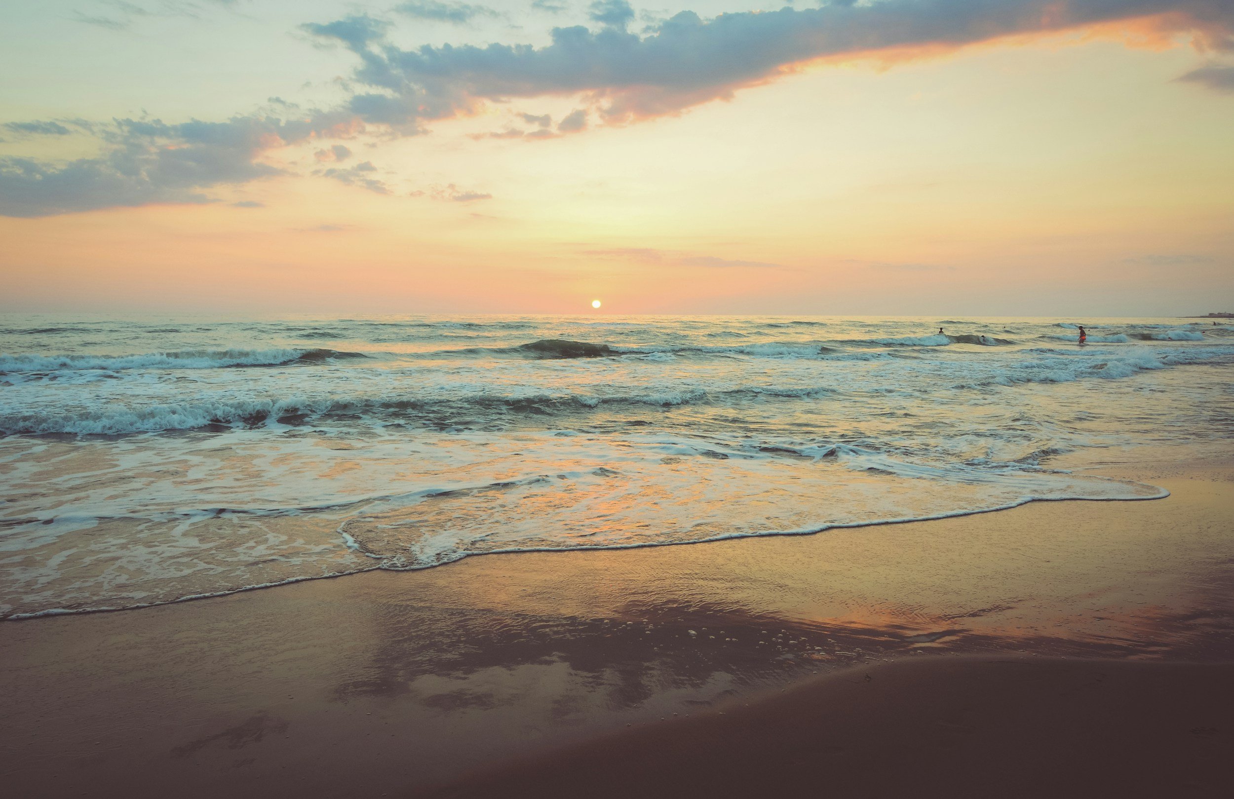 Sunset over the ocean with gentle waves and a partly cloudy sky, sandy beach in the foreground, and a few people in the water.