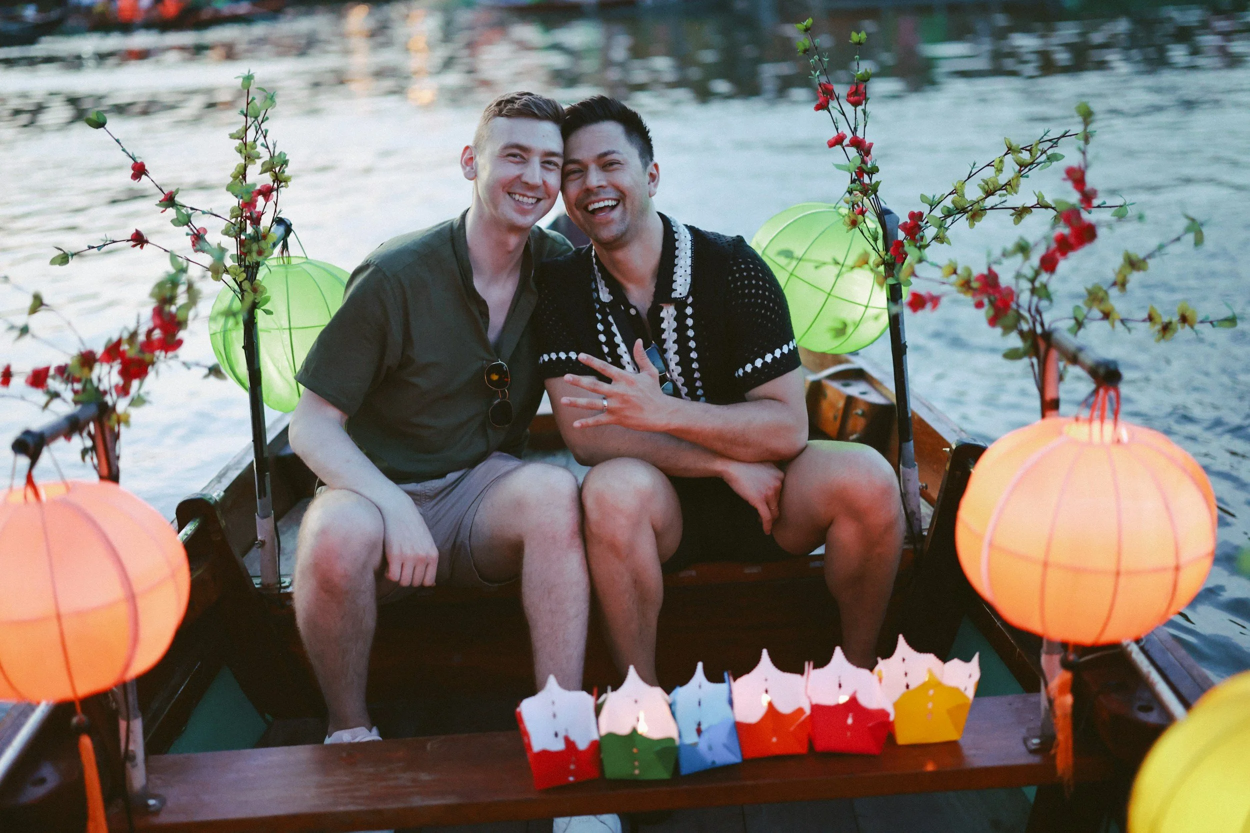 Two men sitting closely together on a decorated boat with lanterns and flowers, smiling by a river during sunset.