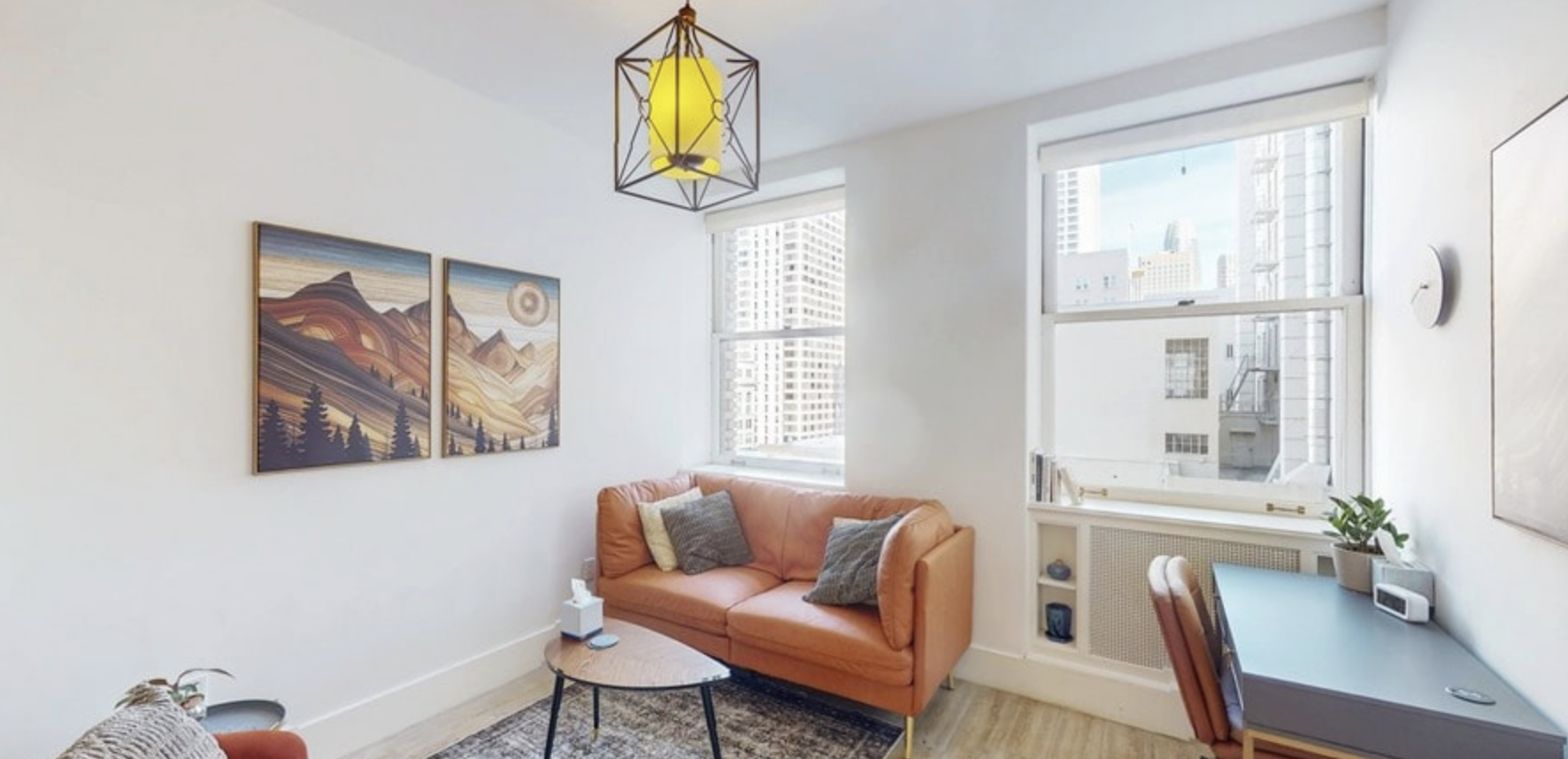 Bright living room with large windows, peach sofa with cushions, wall art depicting mountains and a sun, modern chandelier, side table, desk with a plant and clock.