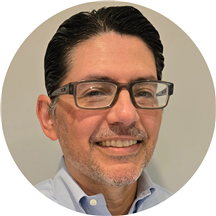 Close-up of a middle-aged man with dark hair, glasses, and a slight beard, smiling in front of a plain background.