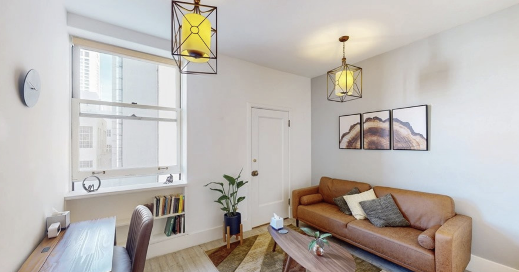 Living room with white walls, a large window, a brown leather sofa with pillows, a wooden coffee table with a small plant, artwork on the wall, a potted plant, a bookshelf, a desk, and hanging yellow pendant lights.