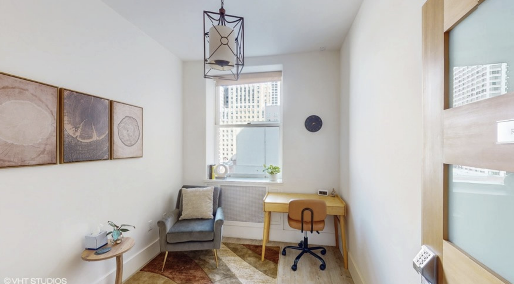 Small room with a gray armchair with beige pillow next to a round wooden side table holding a plant, tissue box, and remote, a window with city view and a small desk with a brown chair beneath the window, a round wall clock, and wall art with three panels.