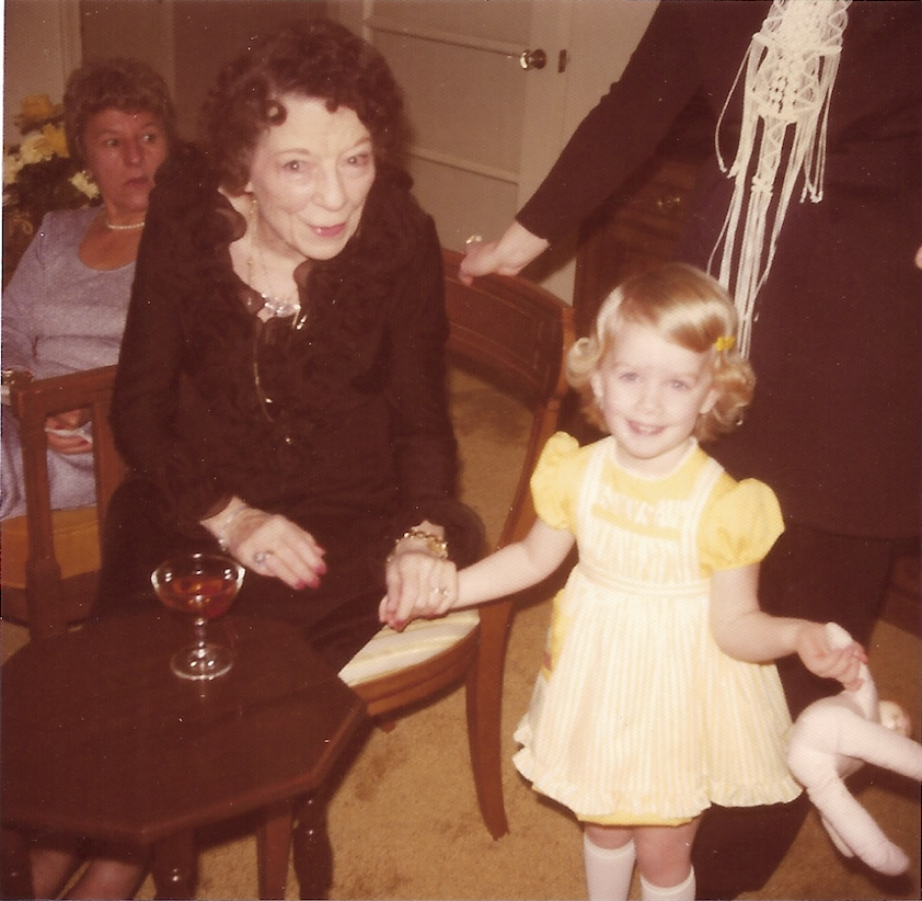 A young girl with blonde hair and a yellow dress holds hands with an older woman with curly dark hair who is seated at a table. The woman is wearing a dark outfit and jewelry, and there is a drink glass on the table. Another woman with dark hair is seated in the background, looking surprised. Part of a person's arm and hand are visible on the right side of the image.