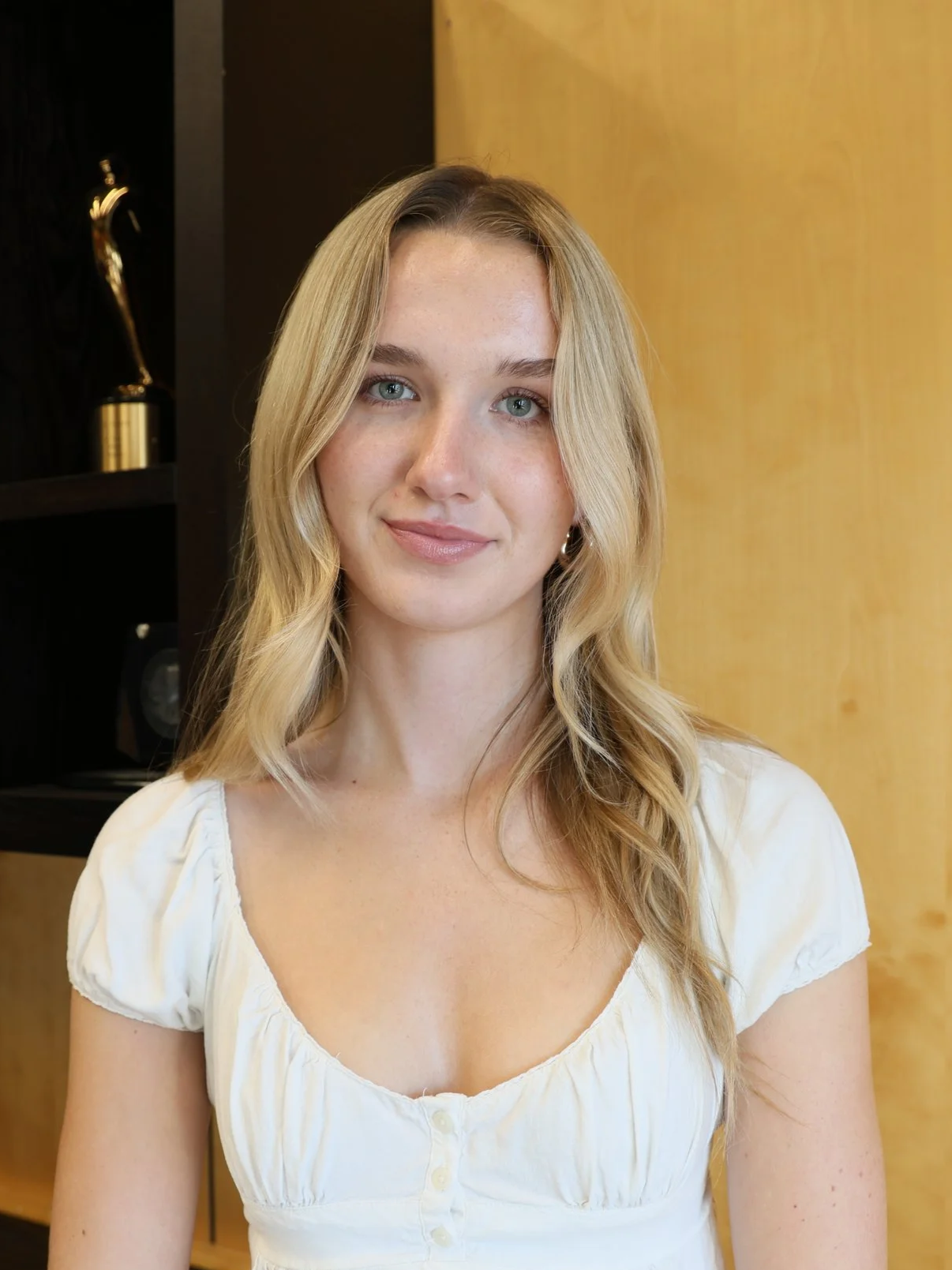 Young woman with long, curly brown hair wearing a cream shirt smiling softly against a set of stairs behind her.