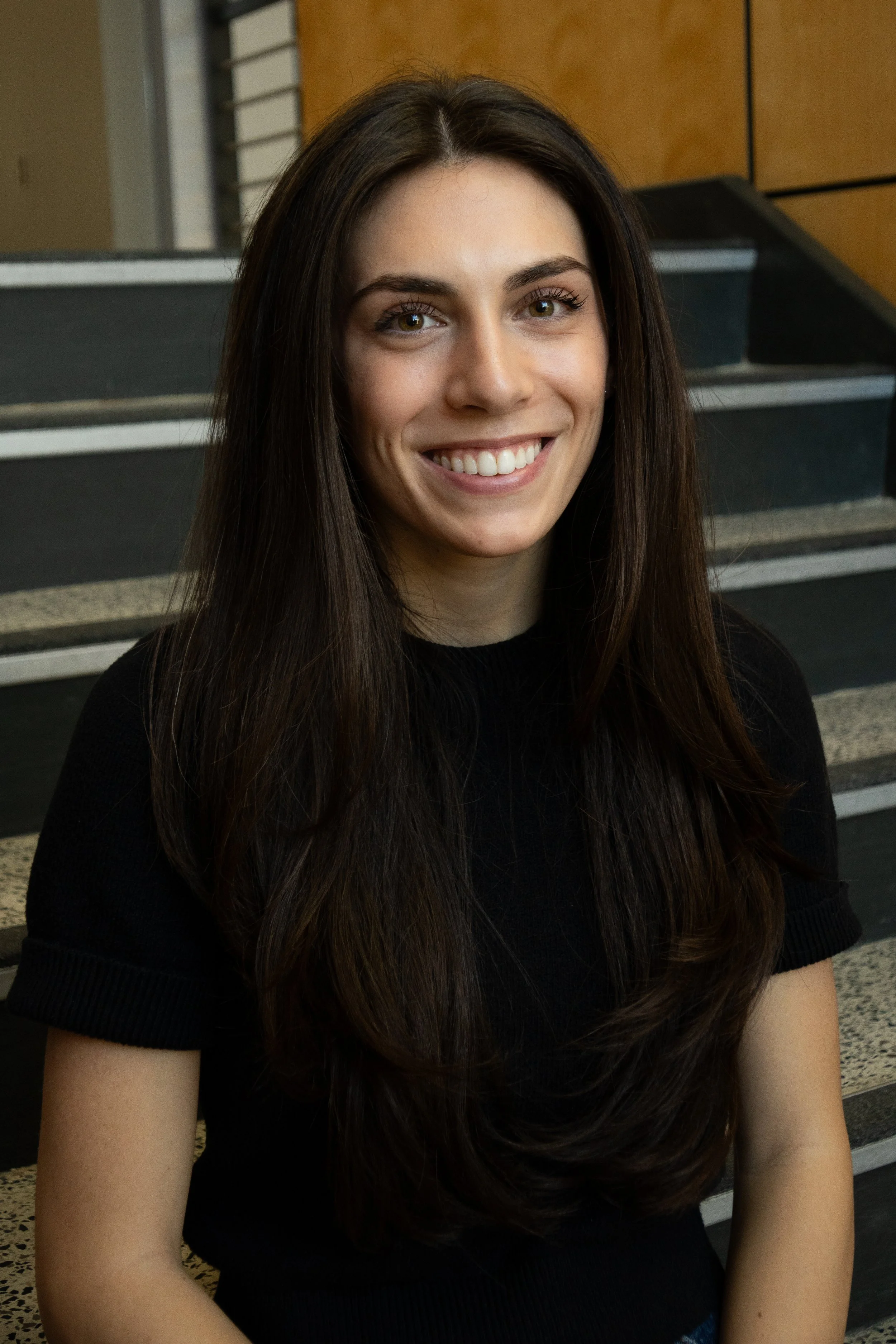 A young woman with long brown hair, smiling, wearing gold jewelry and a black shirt, against a purple background.