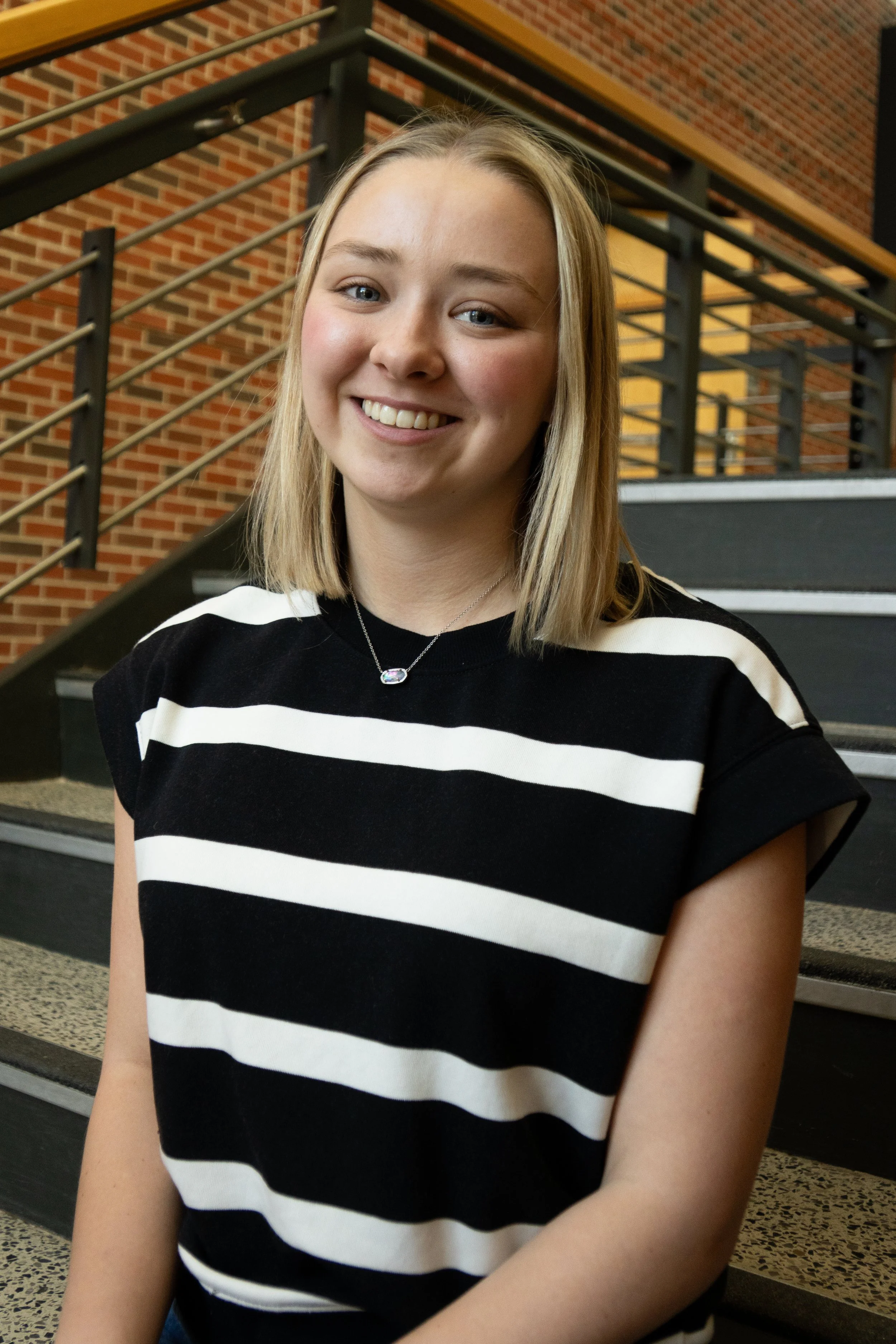 Portrait of a smiling young woman with long blonde hair, wearing a black turtleneck, against a purple background.