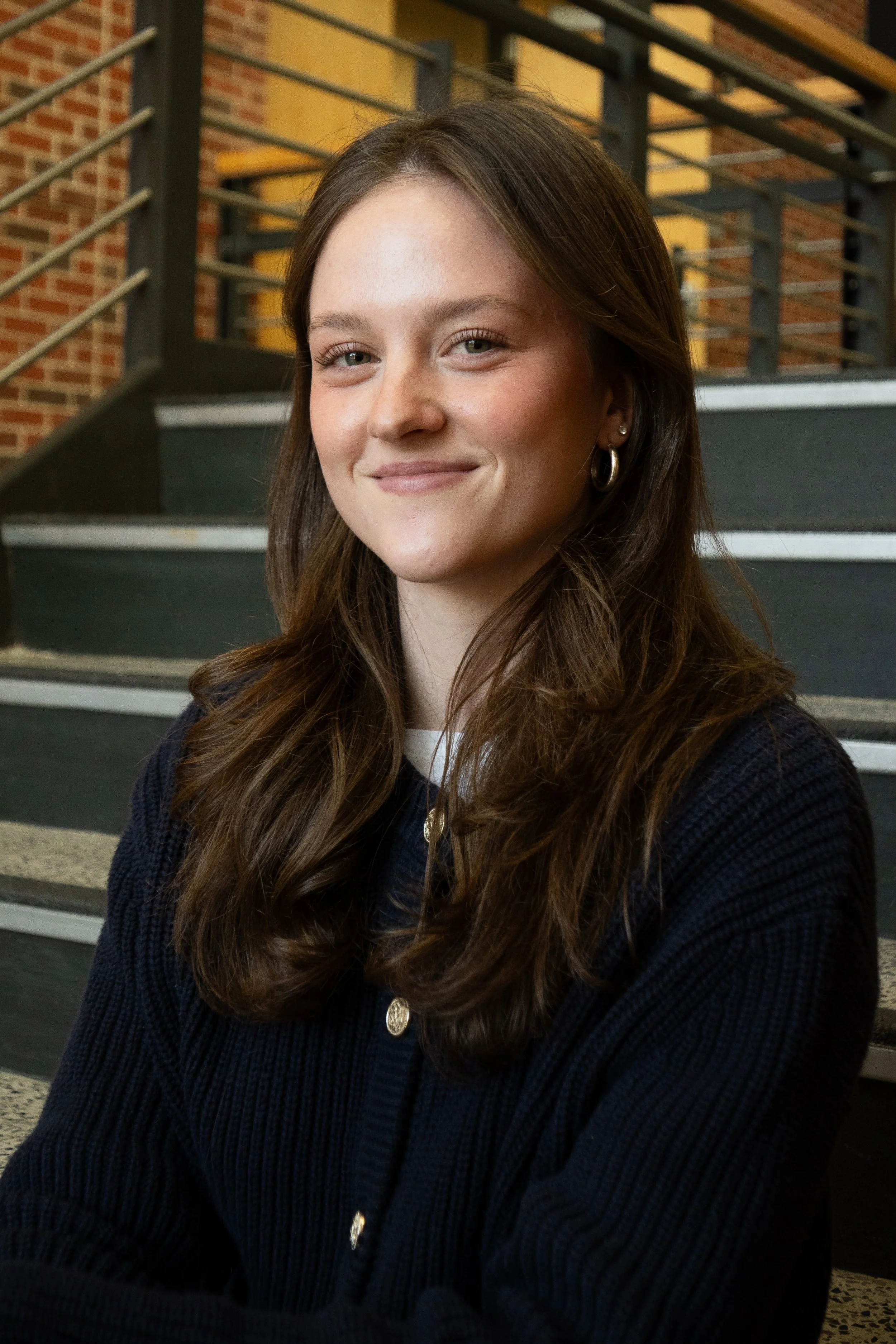 A portrait of a young woman with long brown hair smiling in front of a set of stairs. 