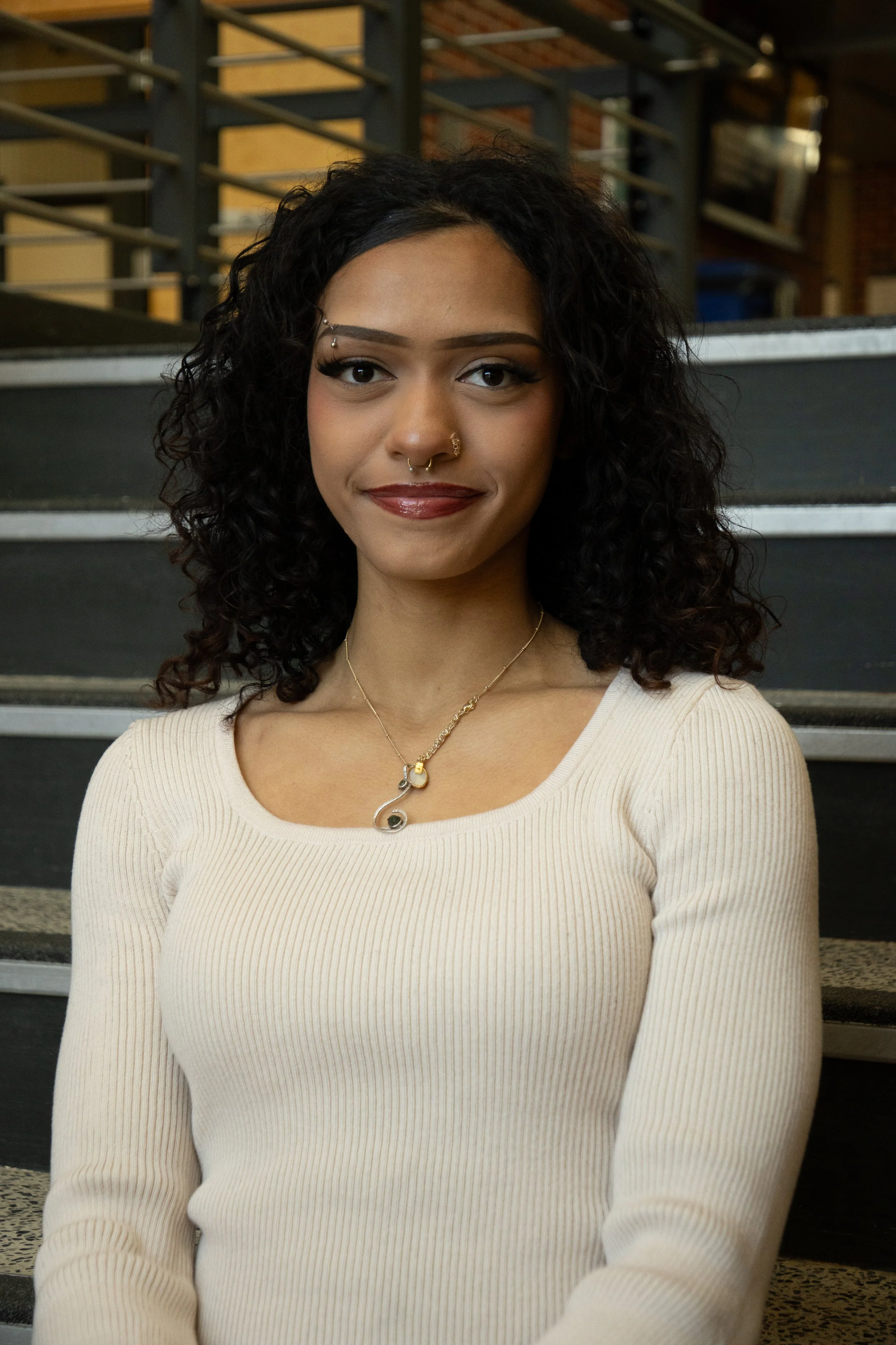Young woman with long, curly brown hair wearing a cream shirt smiling softly against a set of stairs behind her. 