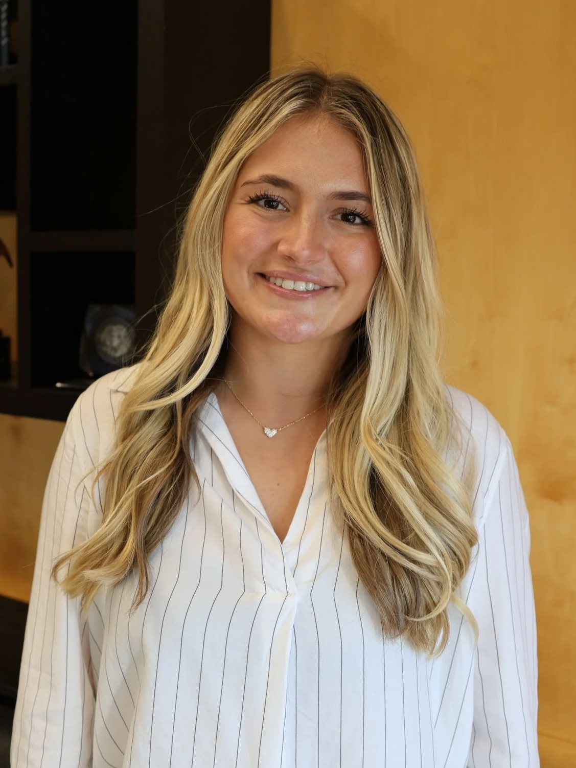 Portrait of a smiling young woman with long blonde hair, wearing a black turtleneck, against a purple background.