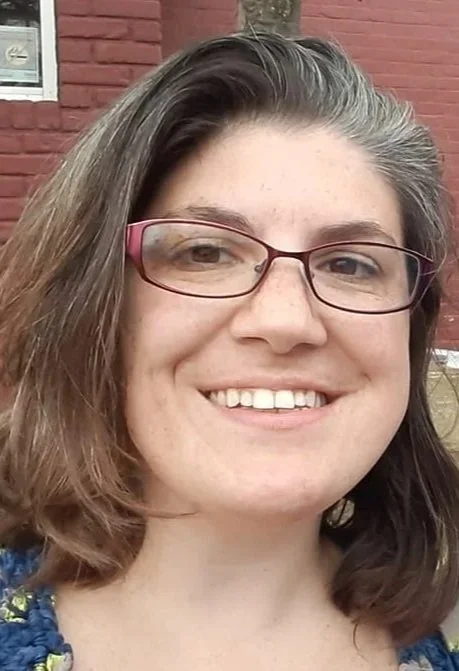 Close-up of a woman with glasses smiling outdoors in front of a red brick building.