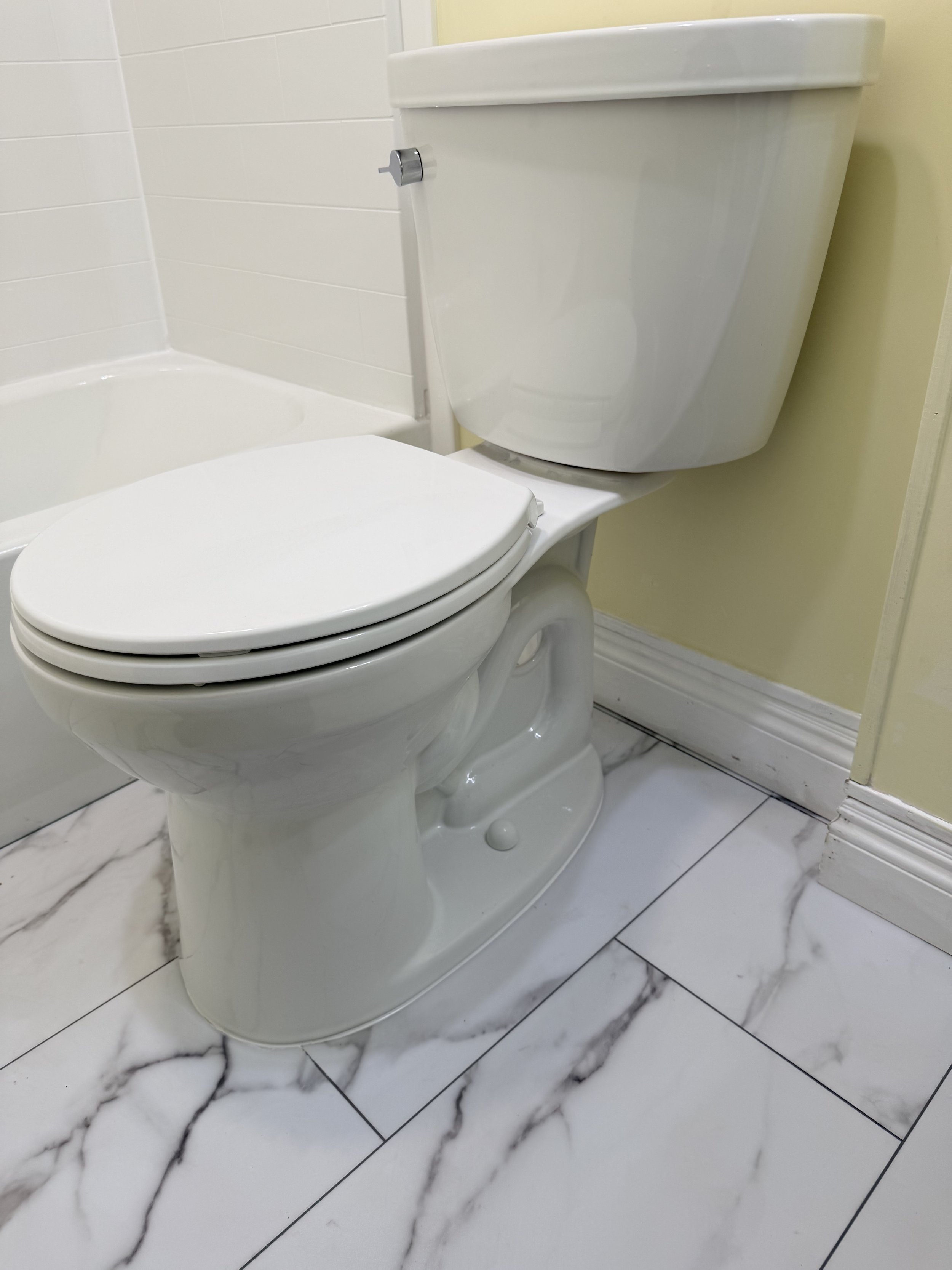 A white toilet next to a bathtub in a bathroom with white marble floor tiles and yellow painted wall. Part of a full bathroom remodel