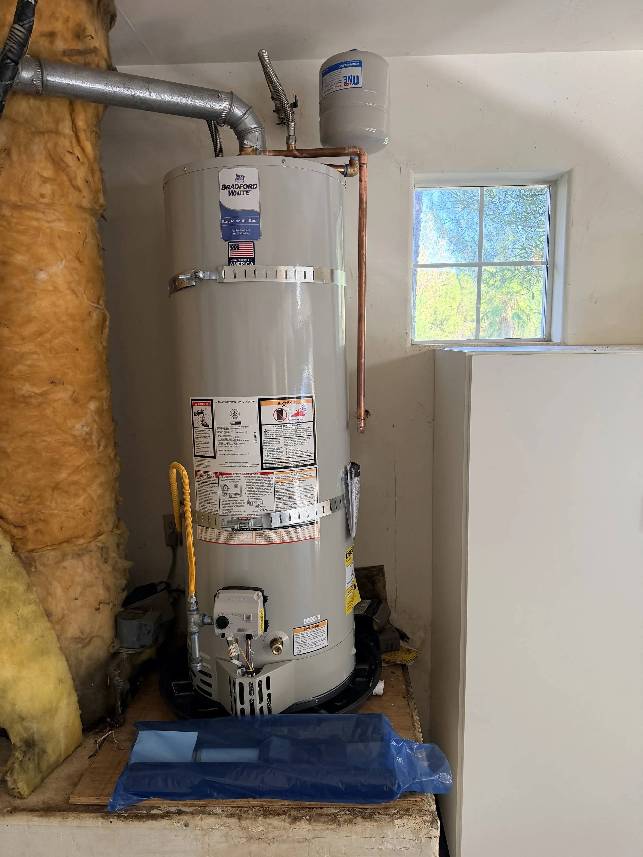 A stainless steel Bradford White water heater installed on a wooden platform in a utility room, with insulation on the left, a window on the right, and pipes and a pressure tank attached.