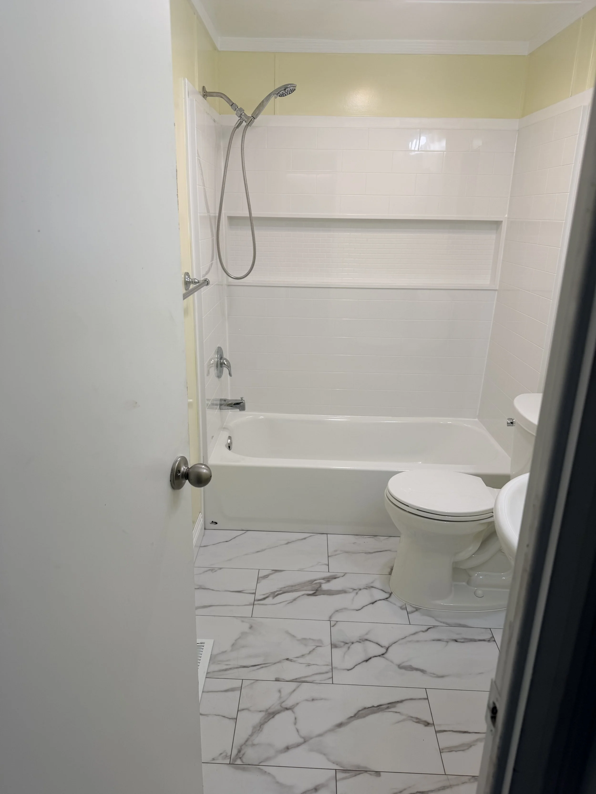 A view of a small bathroom with a bathtub, showerhead, toilet, and marble-style floor tiles.
