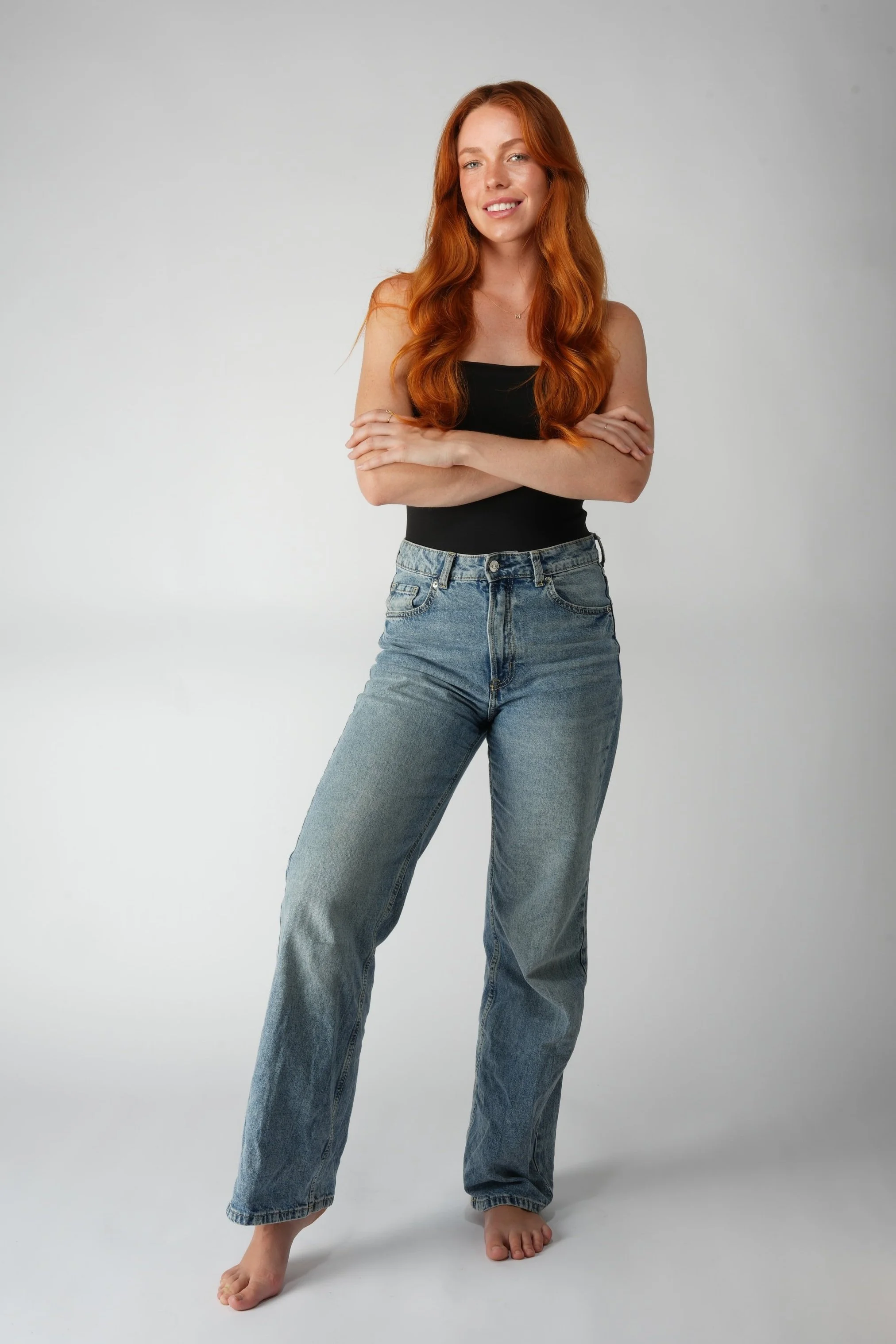 Red-haired woman standing with her arms crossed, smiling, wearing a black tank top and blue jeans in a studio with a plain background.