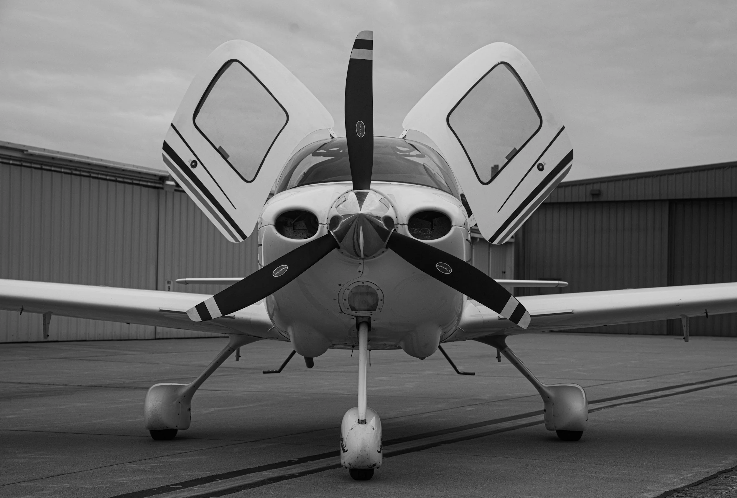 Front view of a small propeller airplane with open cockpit doors, parked on tarmac with hangar in background in black and white.
