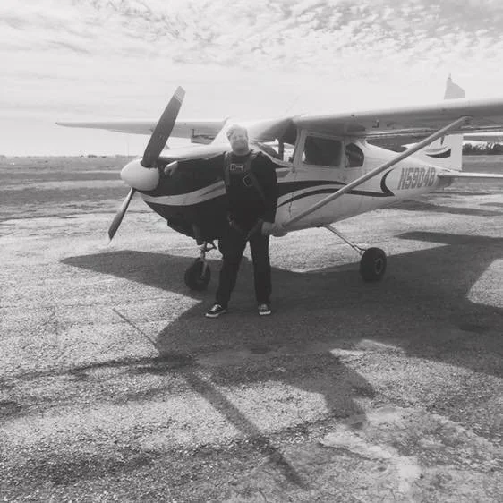Person standing in front of a small single-engine airplane on the tarmac, with open sky and clouds in the background.