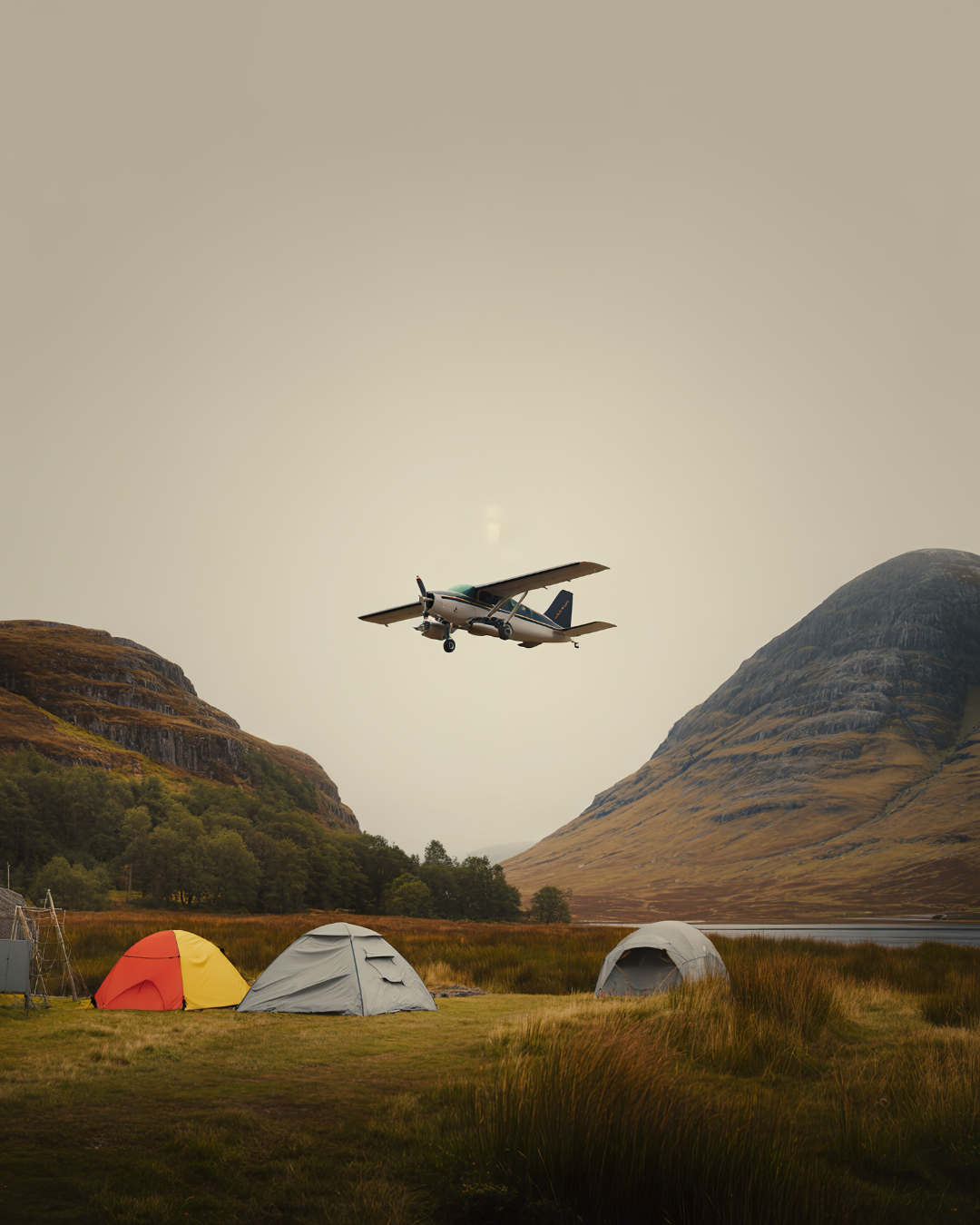 Small airplane flying over a campground with tents, mountains, and a lake, under a cloudy sky.