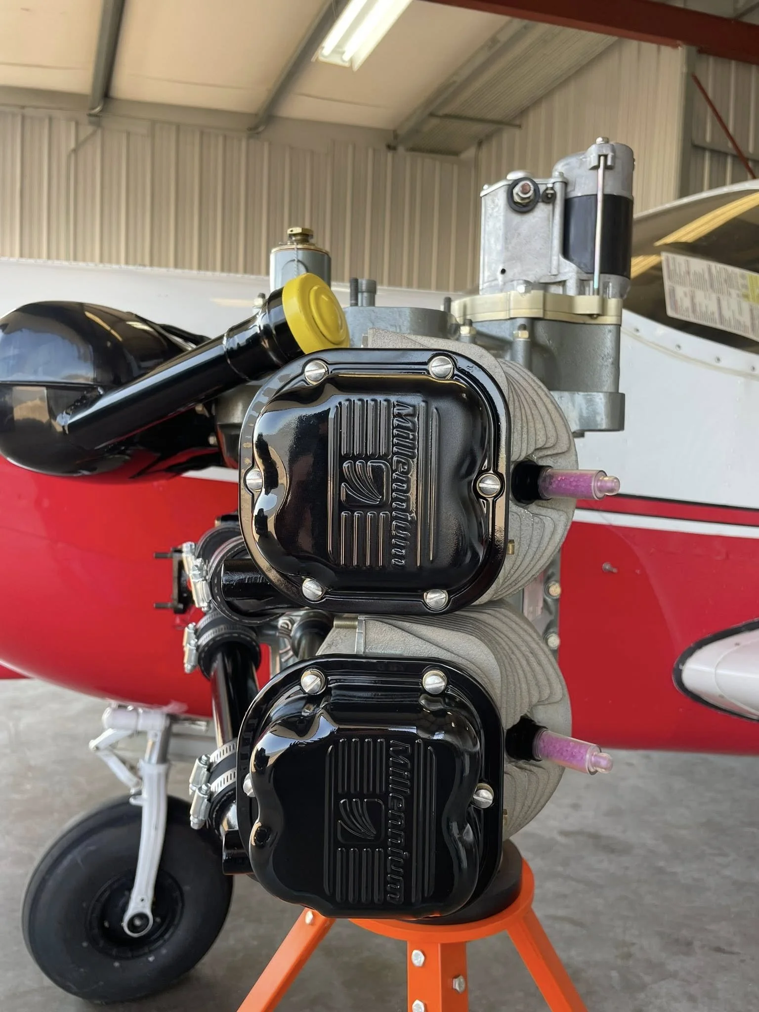 Close-up of a vintage racing motorcycle engine on an orange stand, showing black valve covers with 'Norton' logos, with a garage and aircraft in the background.