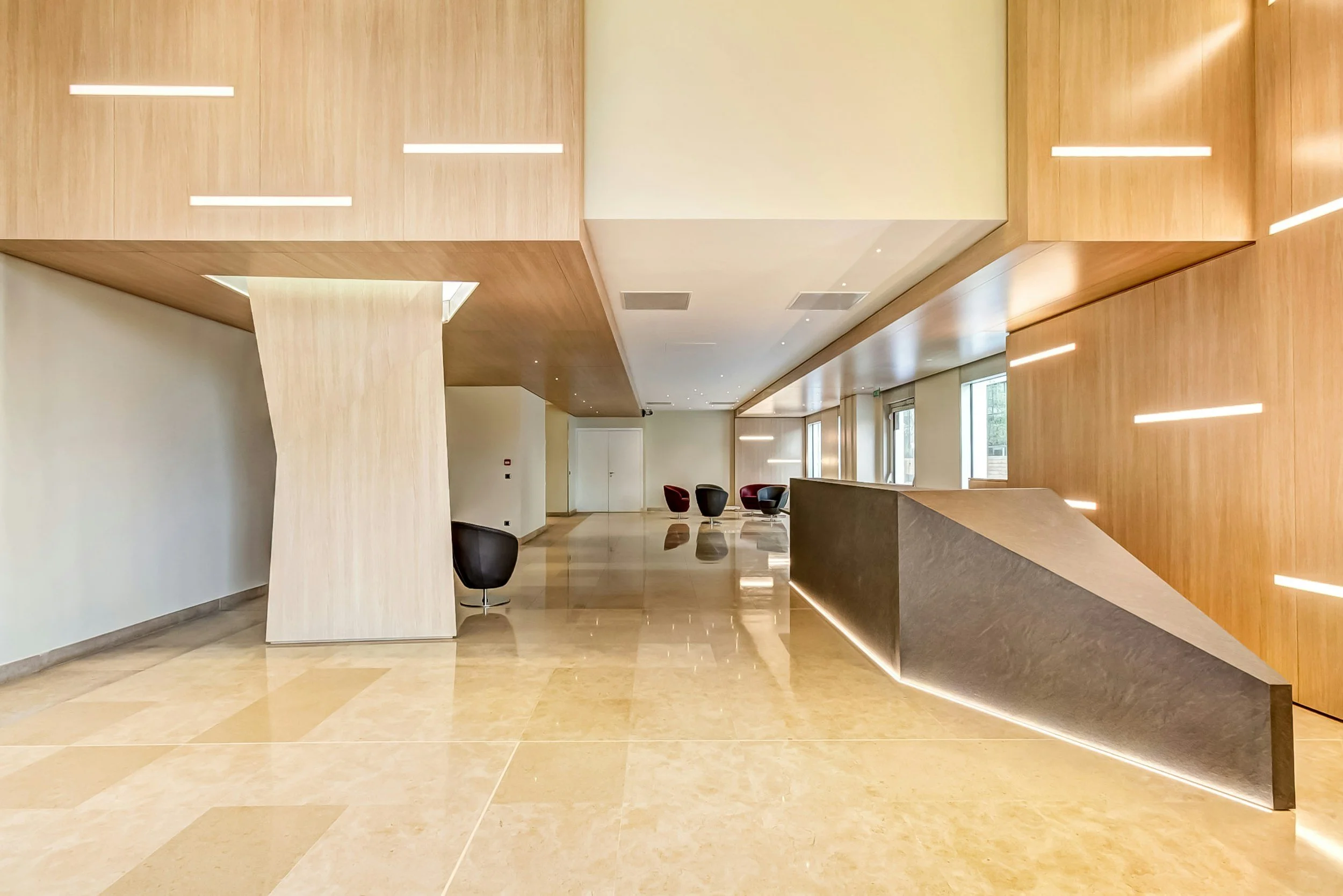 Modern lobby with polished tile floors, wooden wall panels, large windows, and a curved reception desk, with seating area of chairs in the background.