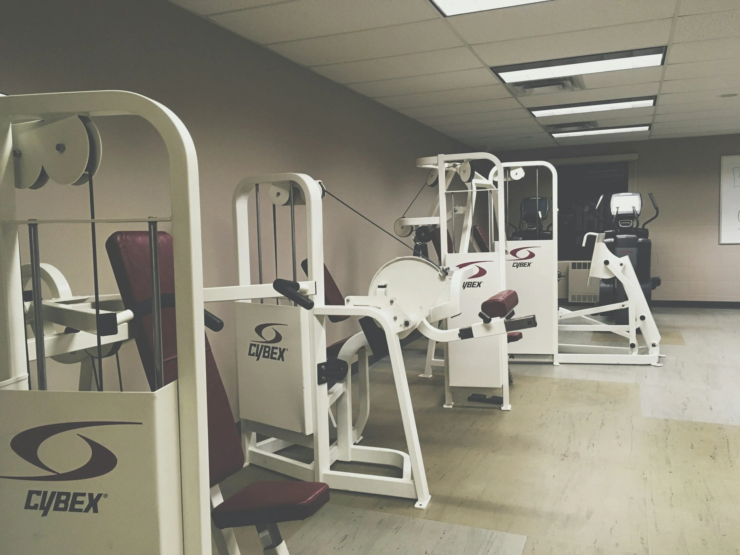 Empty gym room with various white fitness machines and seating, branded Cybex, arranged along the wall with overhead ceiling lights.