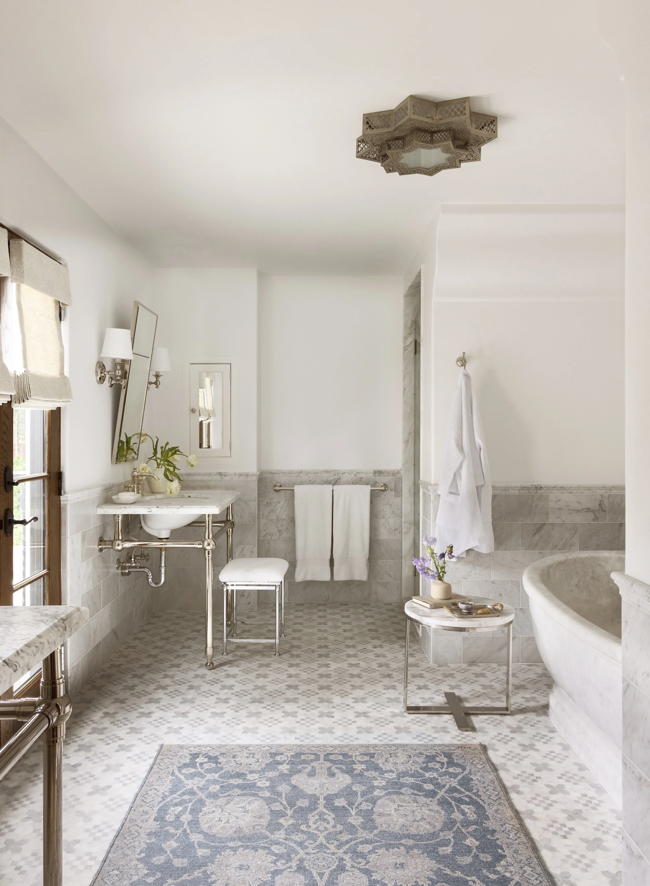Bright bathroom with a white marble sink, a bathtub, a beige patterned rug, potted plants, a ladder towel rack, and a ceiling light fixture.