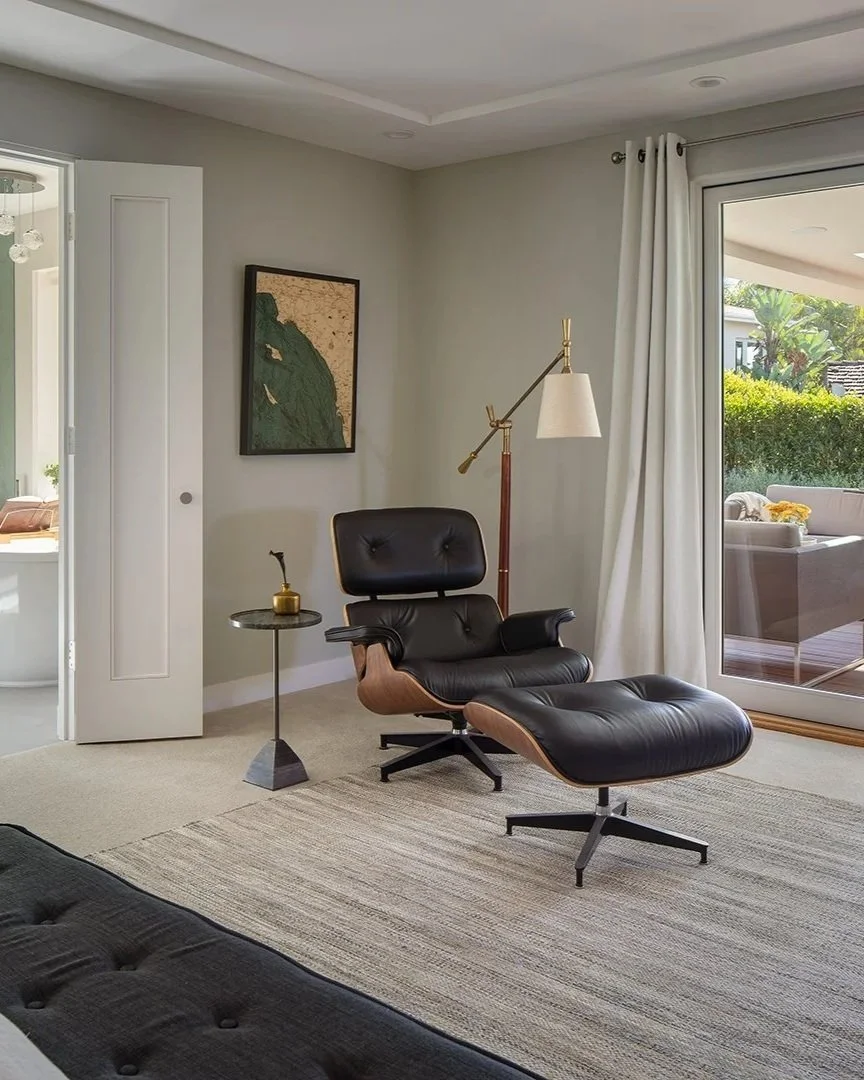 Living room by Tosti Design with a black leather lounge chair and ottoman, a side table with a small plant, a wall art piece, a standing floor lamp, and a sliding glass door leading outside to a patio with greenery and outdoor seating.