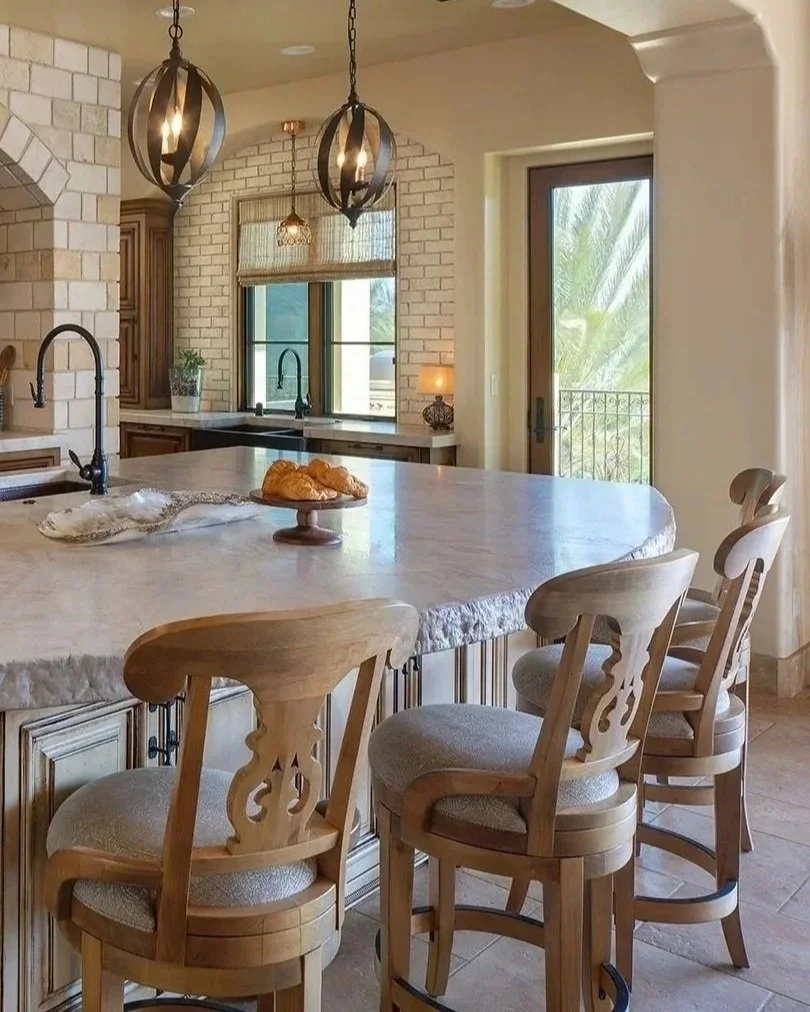 A Del Dios, California kitchen with a large island and three wooden chairs, pendant lights hanging above, a window with blinds, and a door leading outside with a view of palm trees.