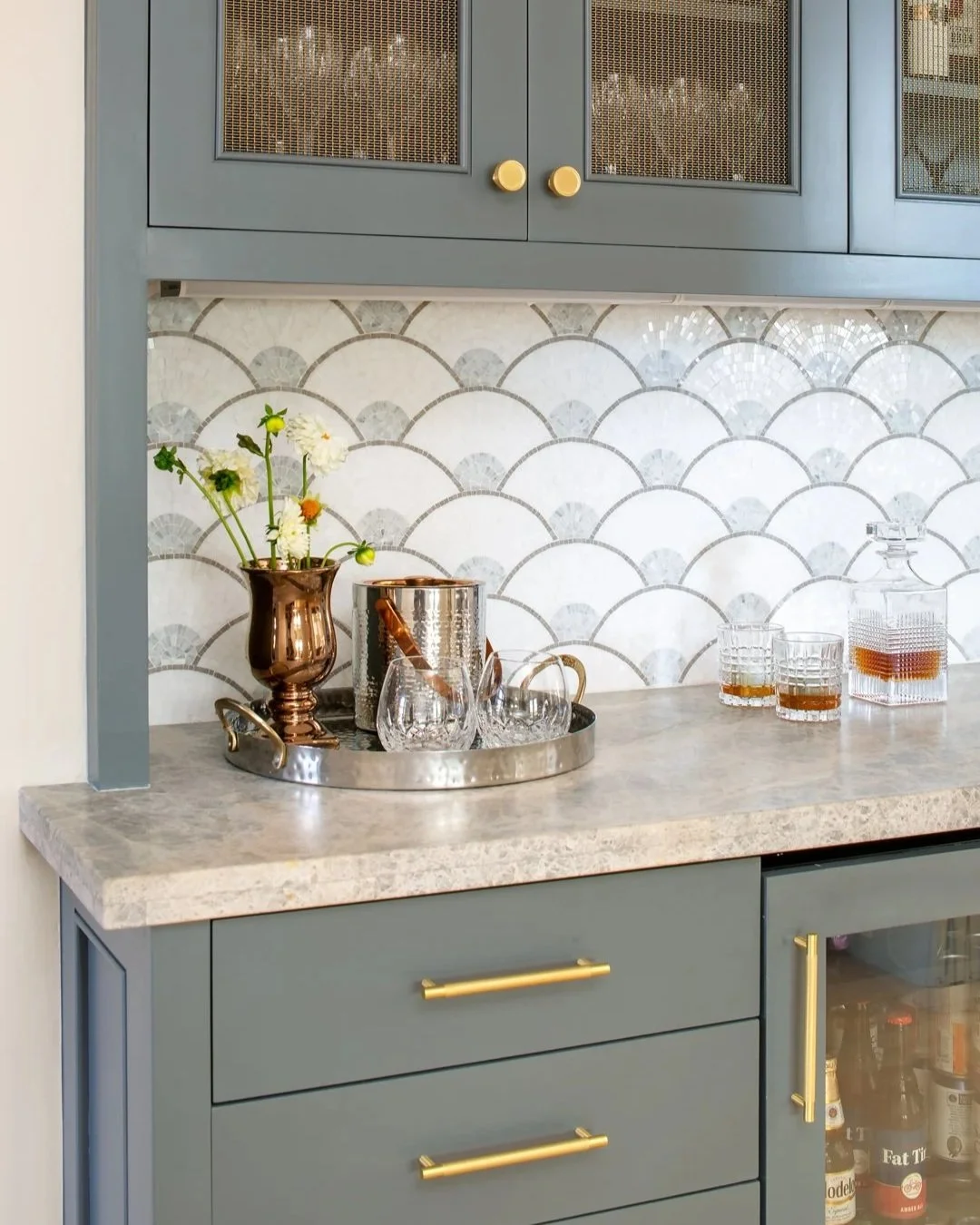 Counter in a kitchen desgined by Tosti Design with a decorative tray holding a vase of flowers, a bottle opener, and glasses, against a background of patterned tile backsplash and blue cabinets with gold handles.