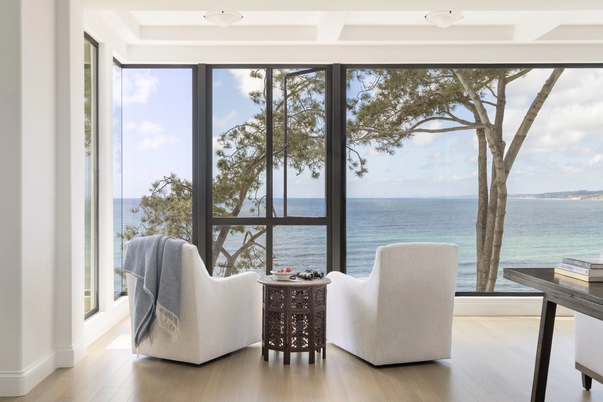 A room with large floor-to-ceiling windows overlooking the ocean, with two white armchairs facing outside, a small ornate table between them, and a blanket draped over one chair. Outside, there are trees and clear blue water.