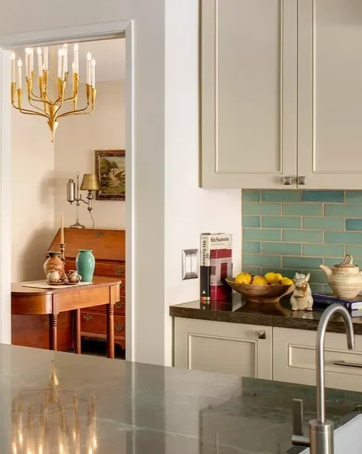 Part of a kitchen with light gray cabinets, a black countertop, and blue subway tile backsplash. On the counter, there is a napkin holder, a bowl of lemons, and a decorative cat figurine. A doorway leads to a dining room with a wooden table, green vase, and a gold chandelier.
