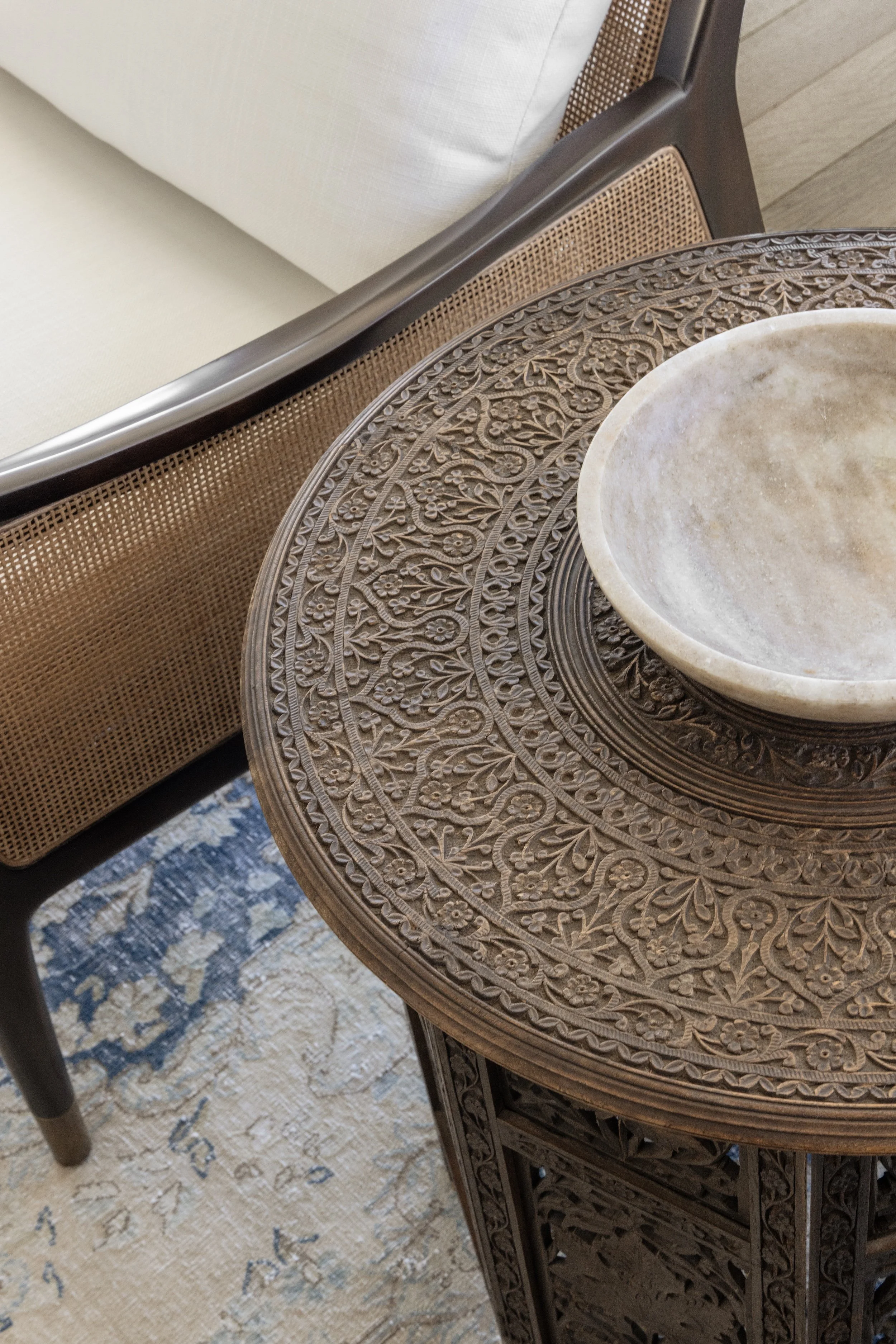 Close-up of a carved wooden side table with a round top, a beige decorative bowl on top, a beige sofa with a rattan back, and a patterned rug on the floor.
