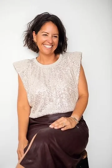 Tosti Design founder and principal Sam Tosti smiling, wearing a sparkly sleeveless top and dark pants, sitting against a plain white background.