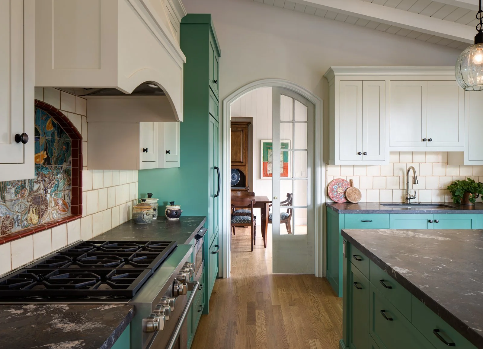 Colorful La Jolla Shores kitchen designed by Tosti Design with white and green cabinets, black countertops, a stove, decorative plates, potted plant, and a glass-paneled door leading to a dining area with wooden chairs and artwork on the wall.