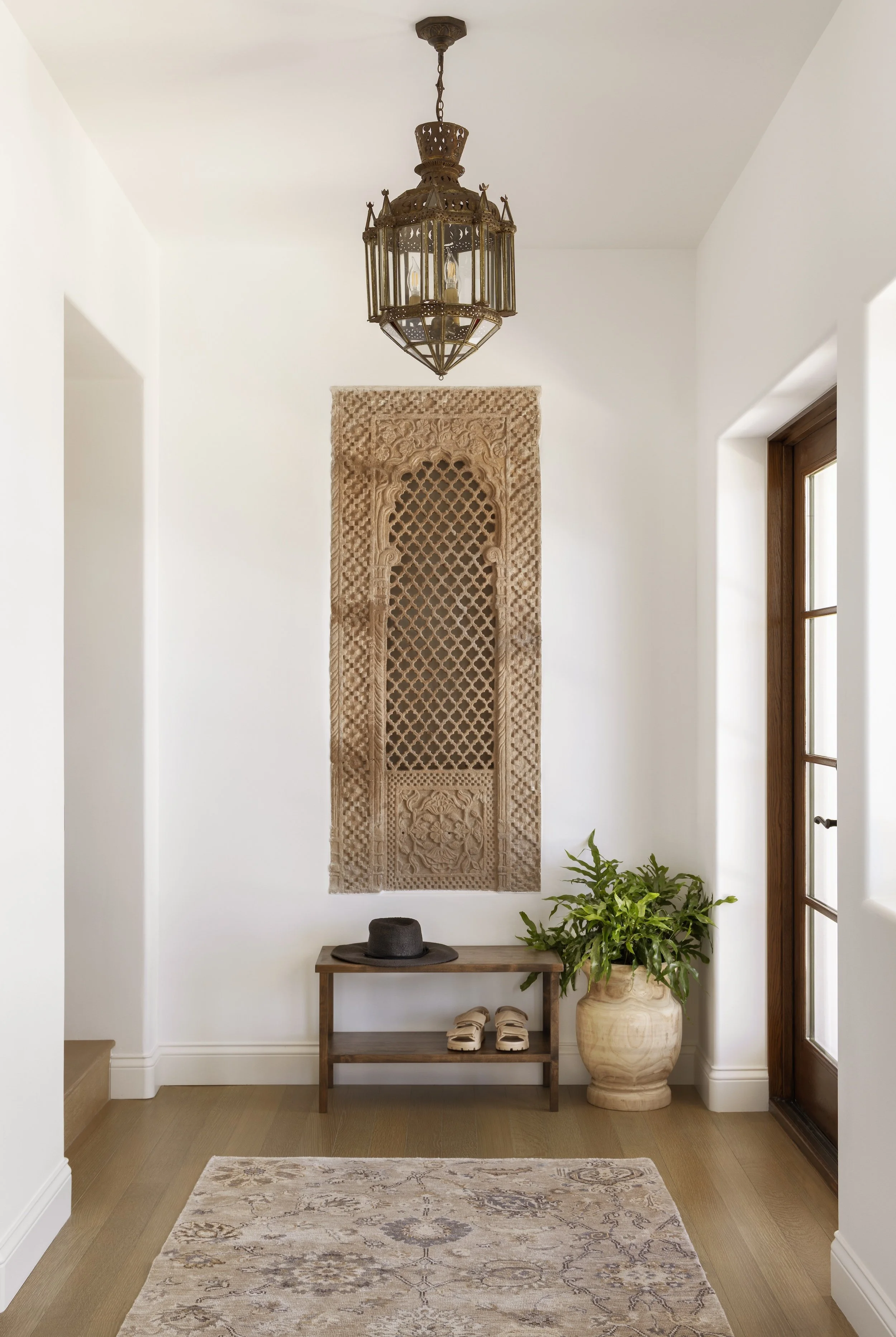 Entryway in a Tosti Design home with white walls, a wooden floor, a woven wall hanging, a small wooden bench with a black hat and white shoes, a potted plant, and a brass chandelier hanging from the ceiling.