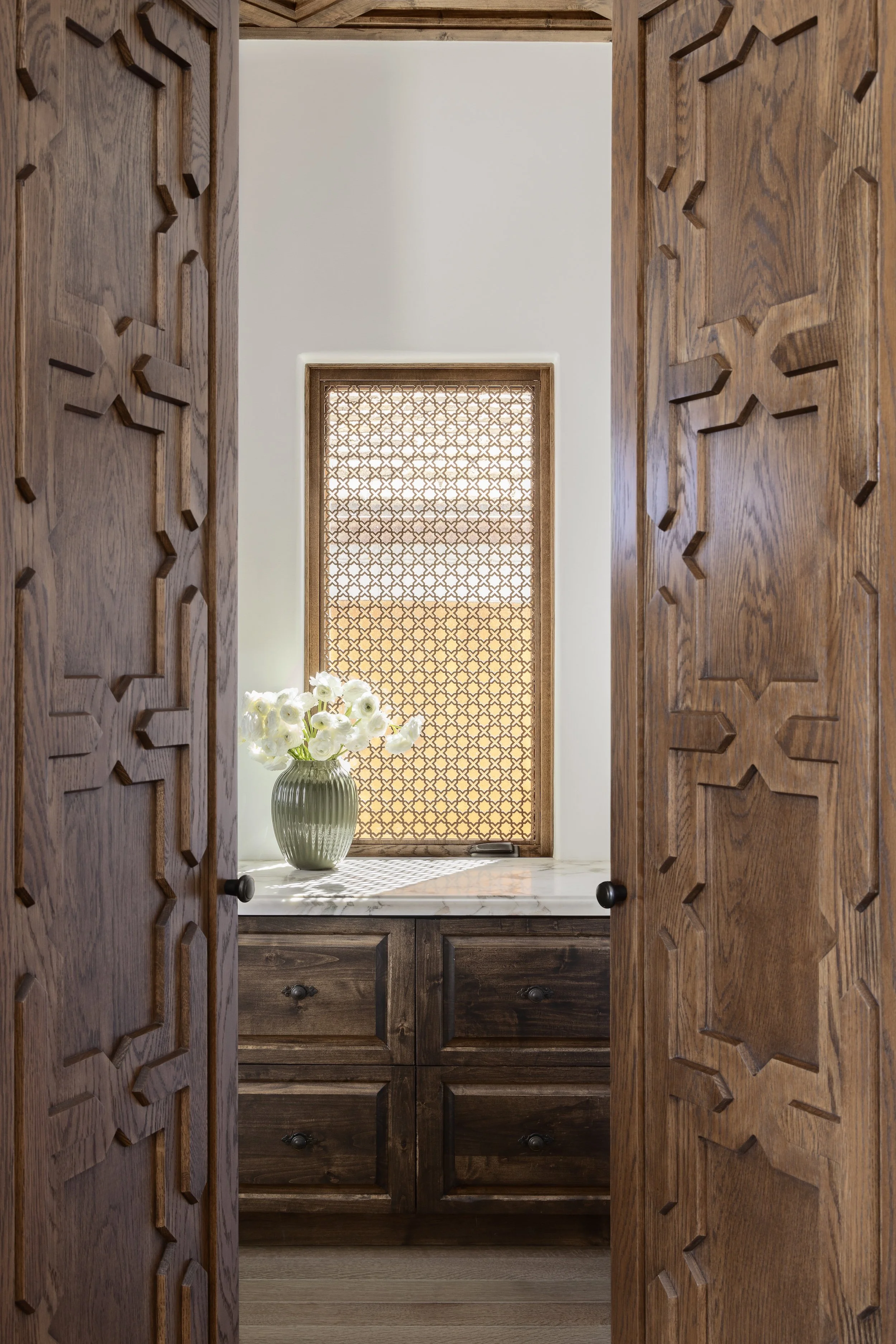 View through carved wooden doors by Tosti Design into a space with a marble countertop, a vase with white flowers, and a window with a patterned wooden screen.