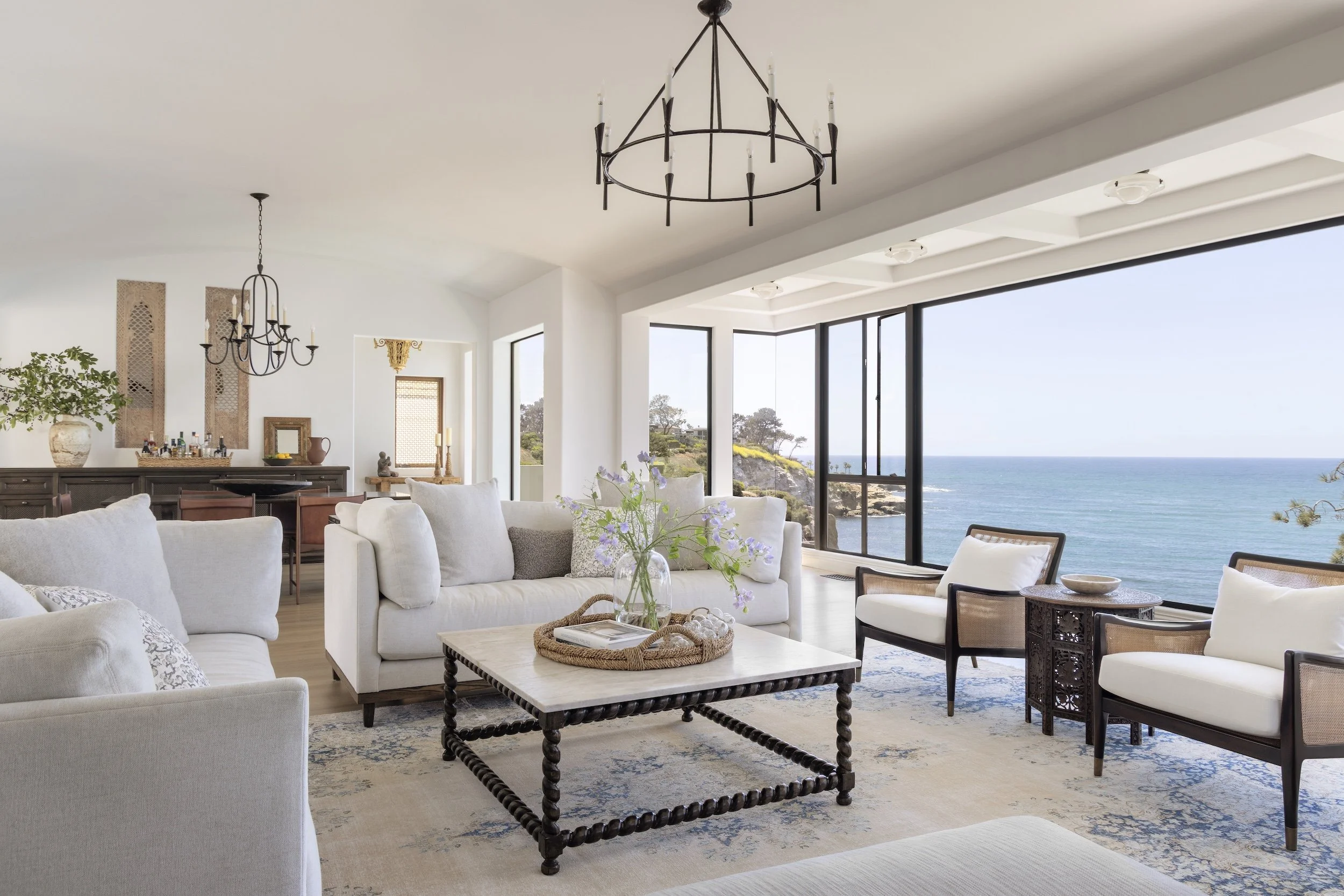 La Jolla Living room designed by Tosti Design with white sofas and chairs, a coffee table with a vase of purple flowers, large windows with ocean view, and chandeliers.