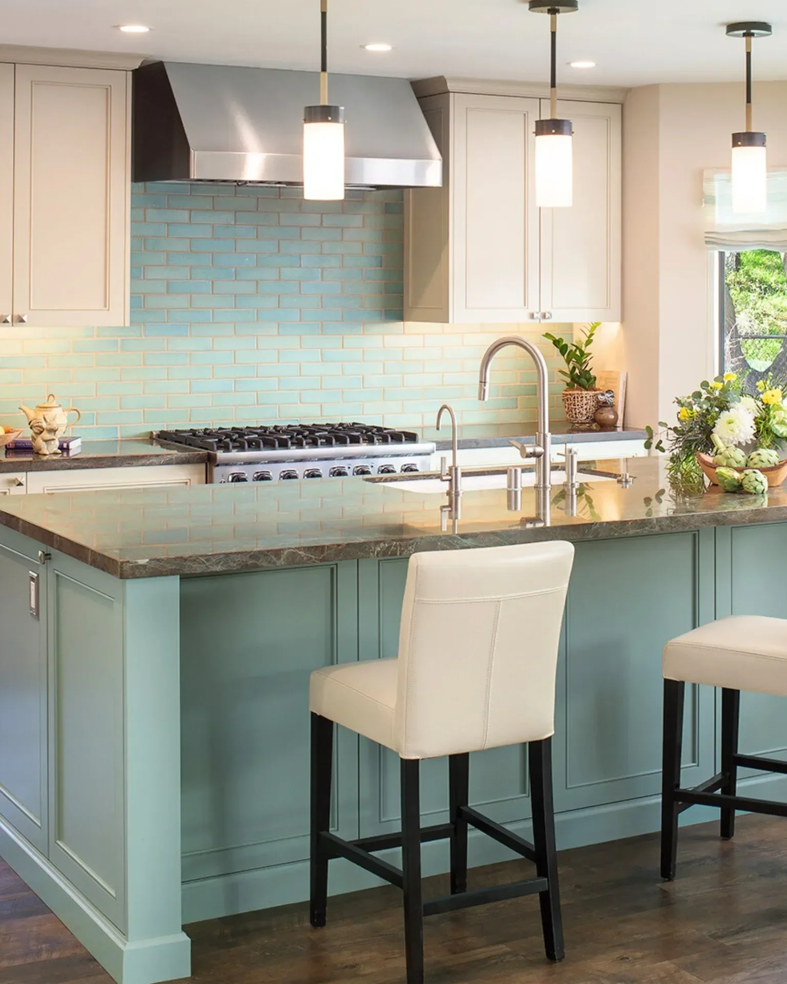 Bear Valley, California kitchen designed by Tosti Design with mint green island, white chairs, beige cabinets, stainless steel stove and range hood, teal brick backsplash, pendant lights, window with view of trees, and decorative plants.