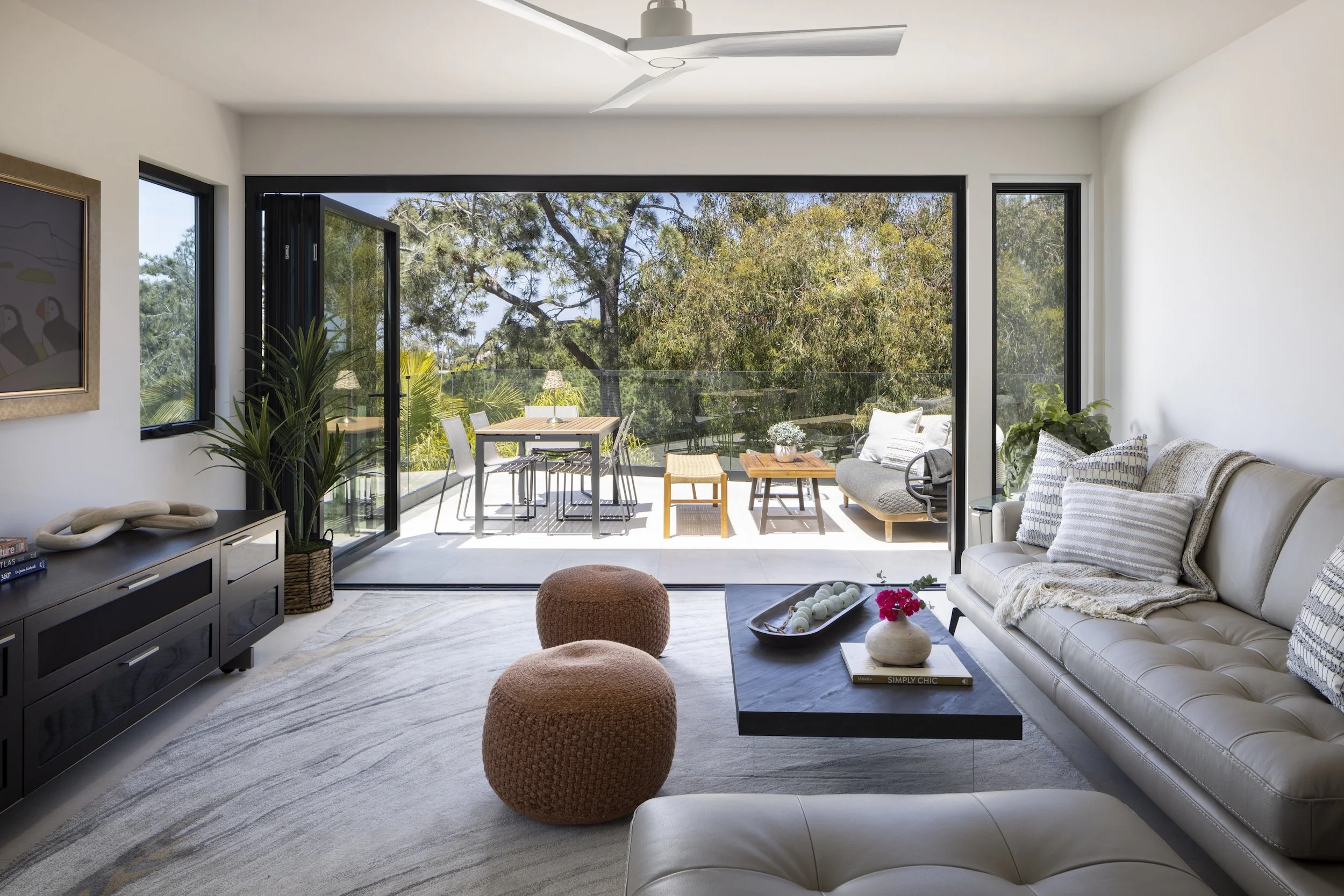 Living room with white walls, a large sliding glass door opening to a balcony with outdoor seating, a gray sofa with patterned pillows, a black coffee table with decorative items, a black TV stand, and various indoor plants.
