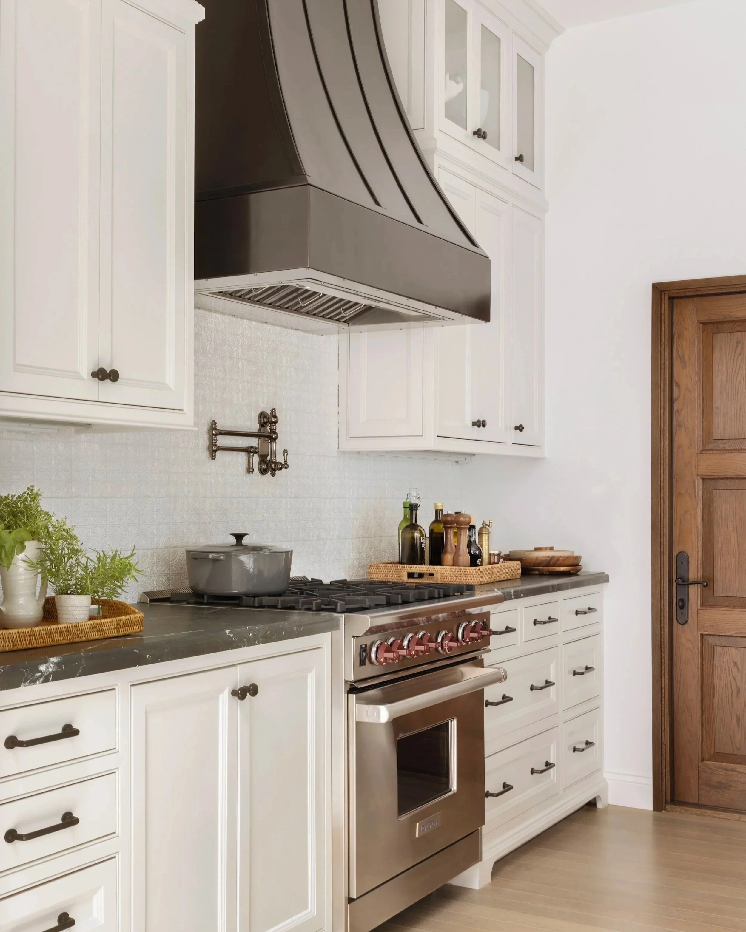 Kitchen designed by Tosti Design with white cabinets, a stainless steel oven, a black range hood, and a wooden door.