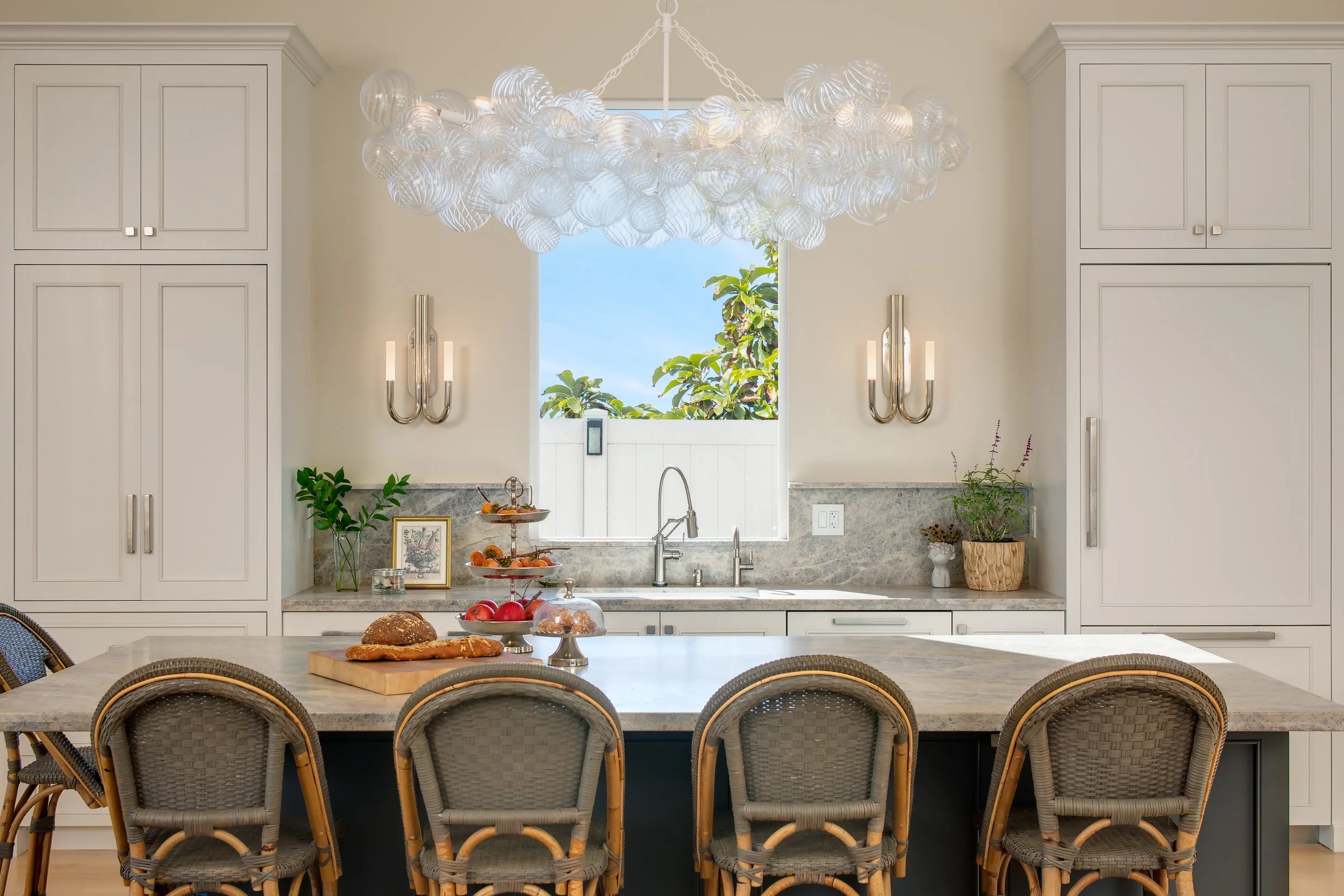 Cardiff, California kitchen designed by Tosti Design with white cabinetry, granite countertops, and a center island with woven rattan chairs. A decorative chandelier hangs from the ceiling.