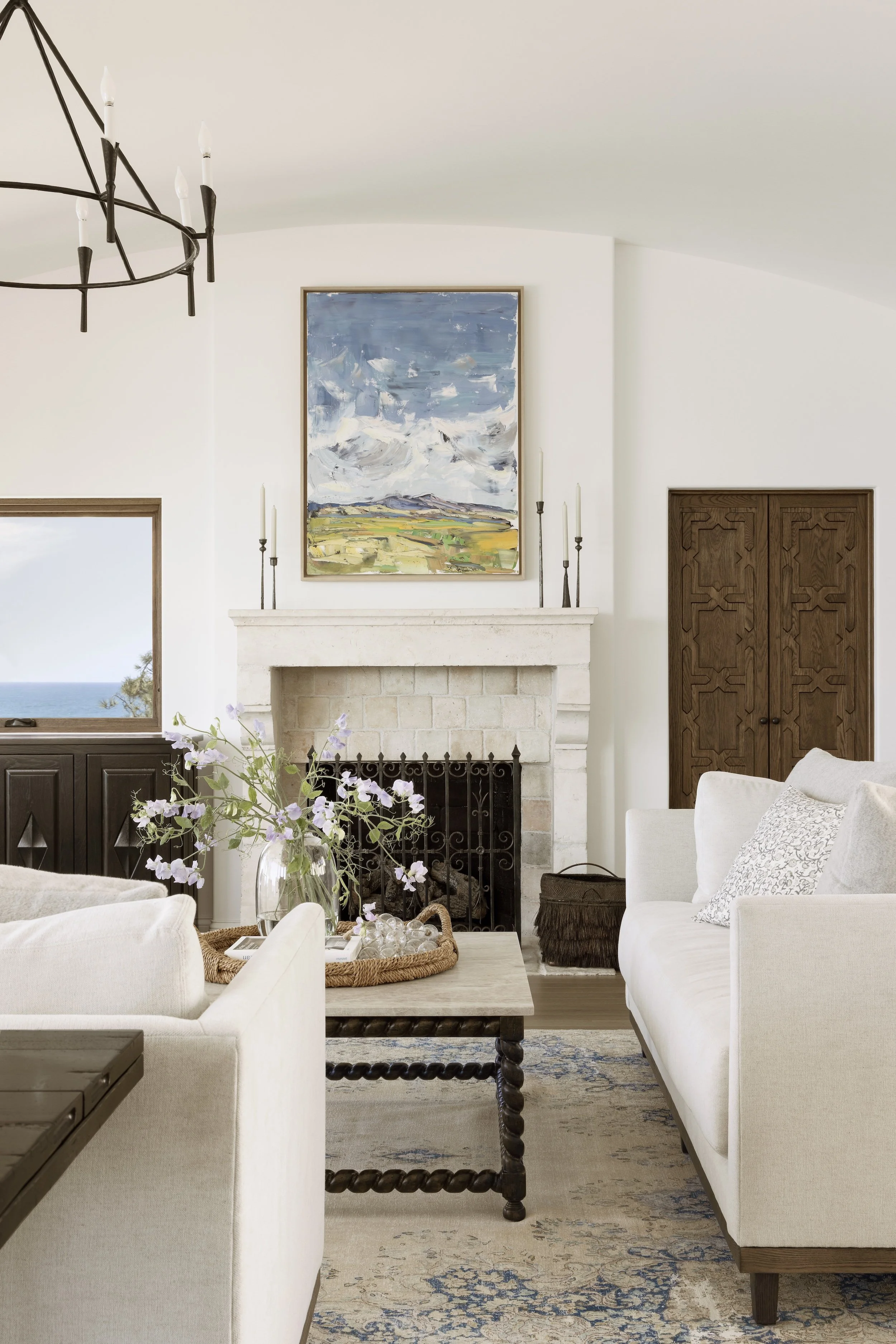 Tosti Design living room with a beige couch, coffee table with a basket and flowers, a fireplace with art above it, and a window showing a view of the ocean.