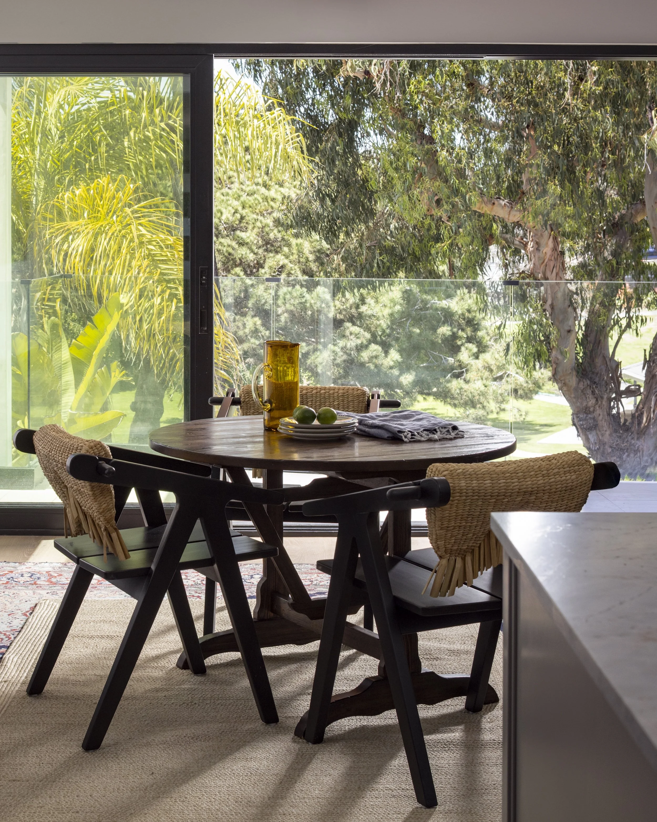Tosti Design dining area with a round wooden table, four chairs with woven and dark wood design, a pitcher and plates with limes, and large glass sliding doors opening to a view of trees and greenery outside.