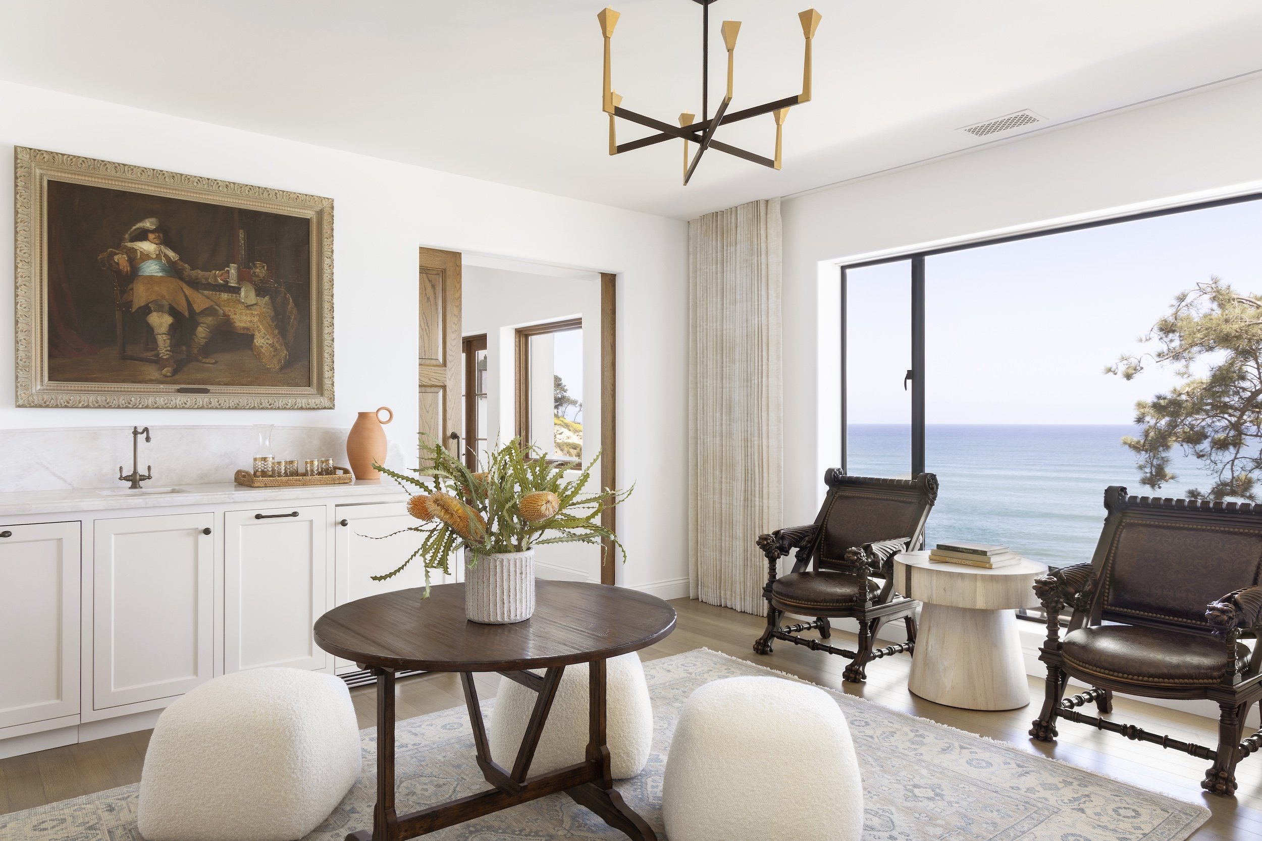 La Jolla Living room by Tosti Desgin with large window overlooking the ocean, dark wooden chairs, a round marble side table, a wooden dining table with a vase of flowers, white cabinets, and wall art.